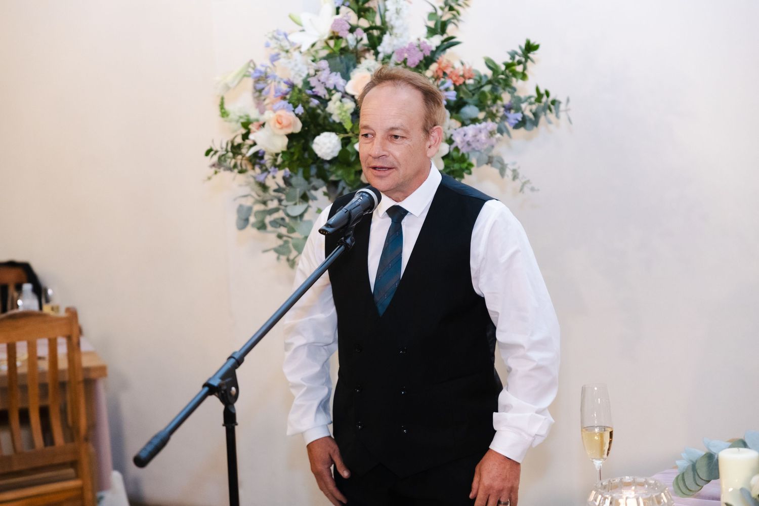 A person in a black vest and tie stands at a microphone with floral arrangements in the background.