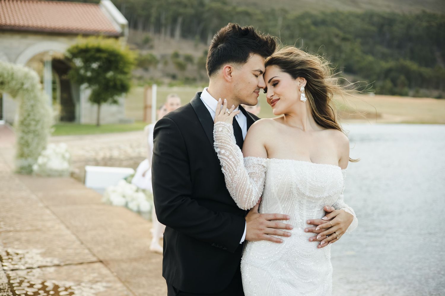 Romantic couple in wedding attire embracing by a lakeside venue, bride in white lace gown and groom in black suit.