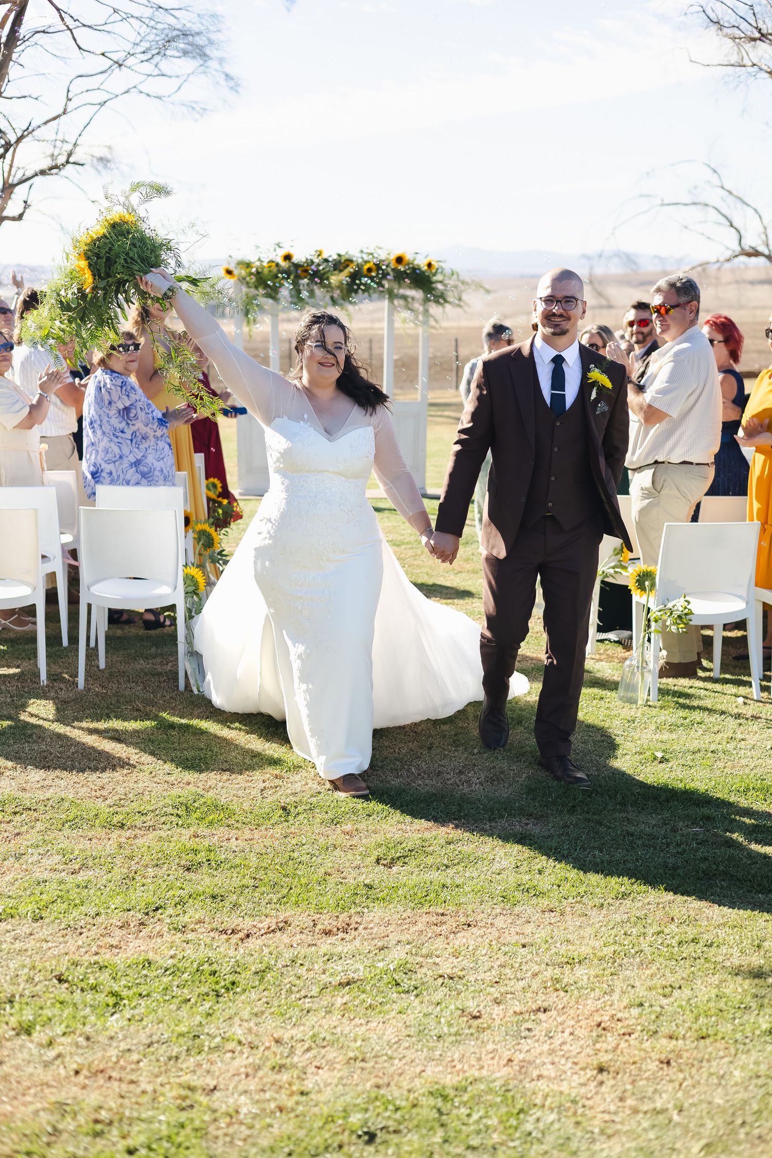 Joyful newlyweds walking down outdoor aisle, bride raising sunflower bouquet in celebration after ceremony.