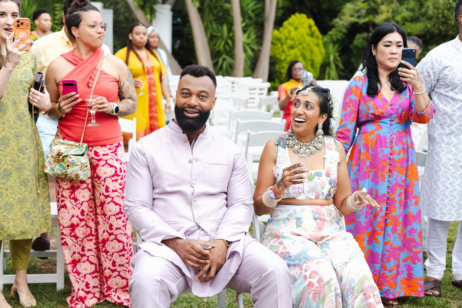 A group of people wearing colorful summer dresses and outfits gather outdoors for a social event in a garden setting.