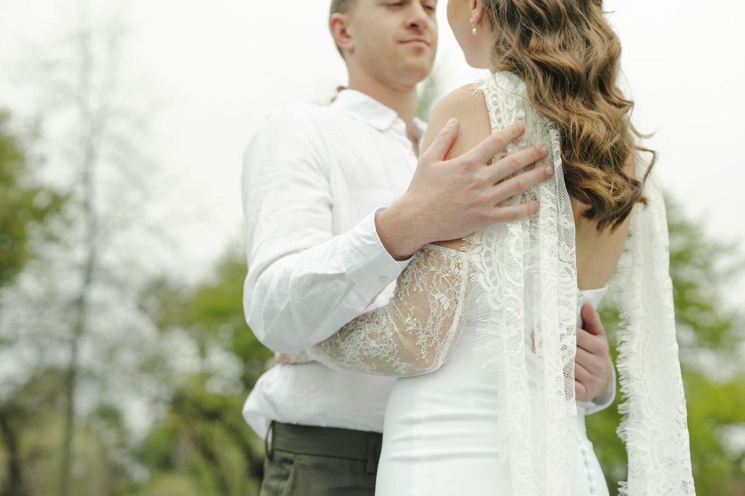 A couple in white wedding attire embraces outdoors, the groom holding the bride close among green trees.