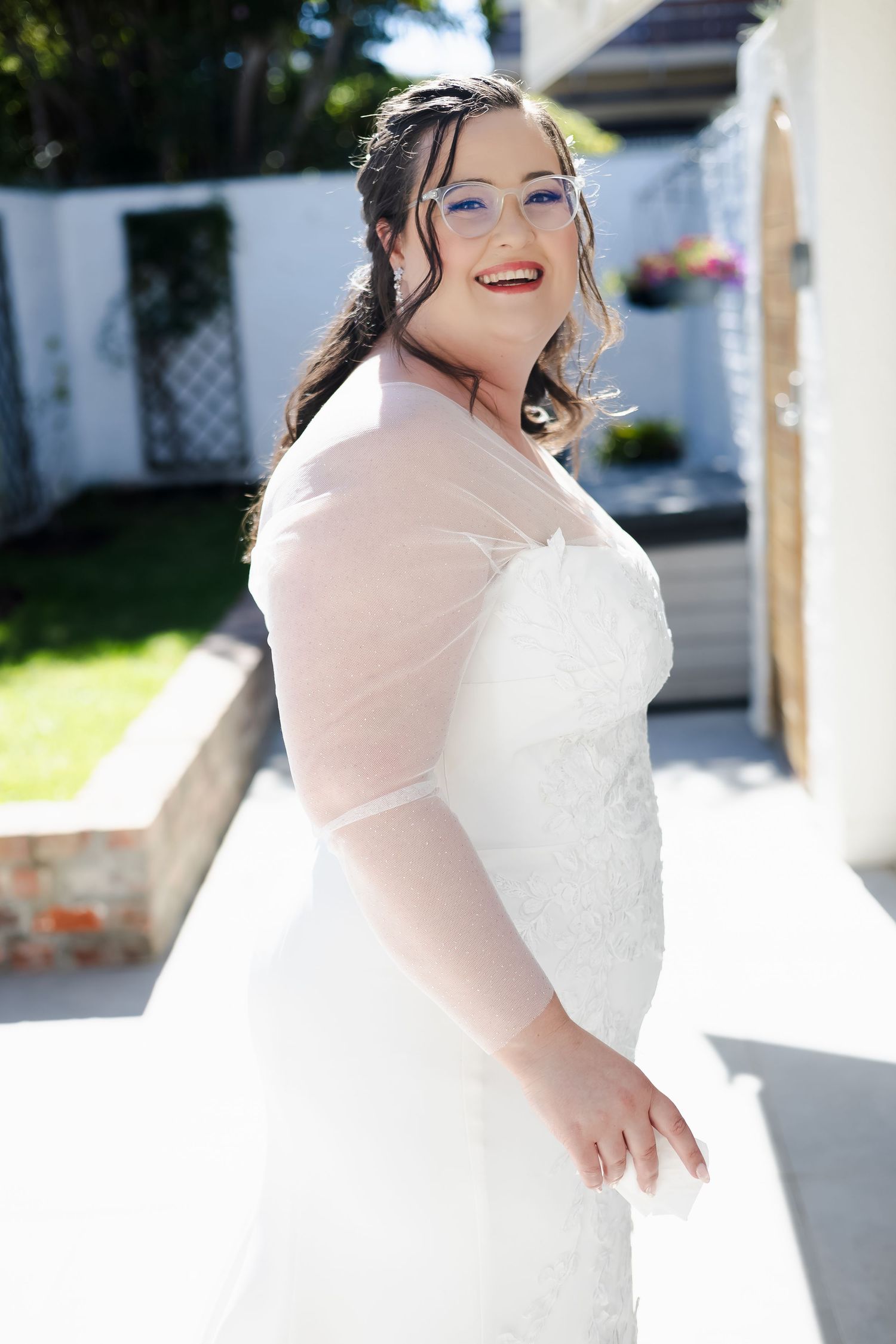 Smiling bride in white lace wedding dress with sheer bolero jacket and blue glasses posing outdoors.