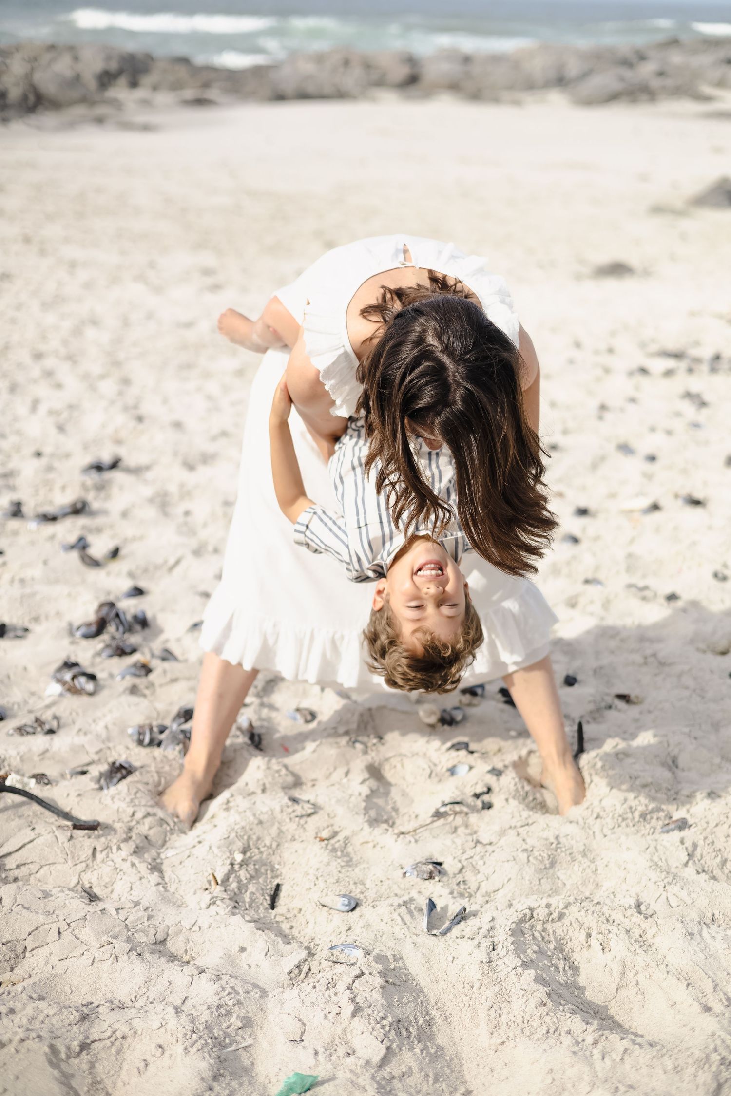 A mother playfully holds her child upside down on a sunny sandy beach, both wearing white outfits.