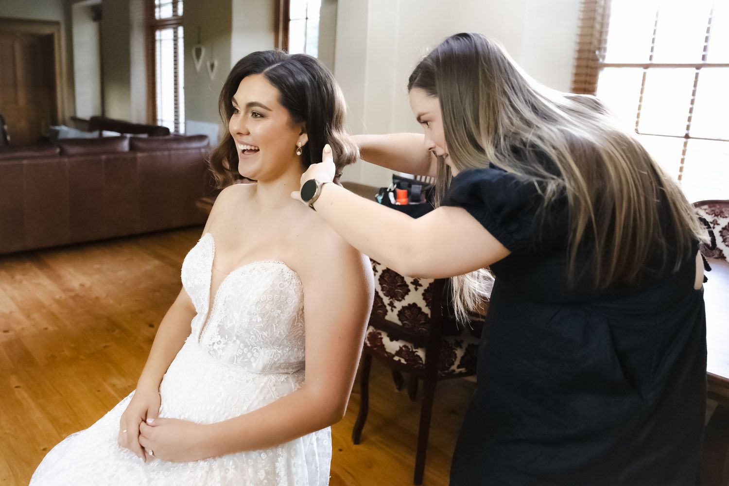 A stylist helps adjust a white lace wedding dress in a room with wooden floors and natural lighting.