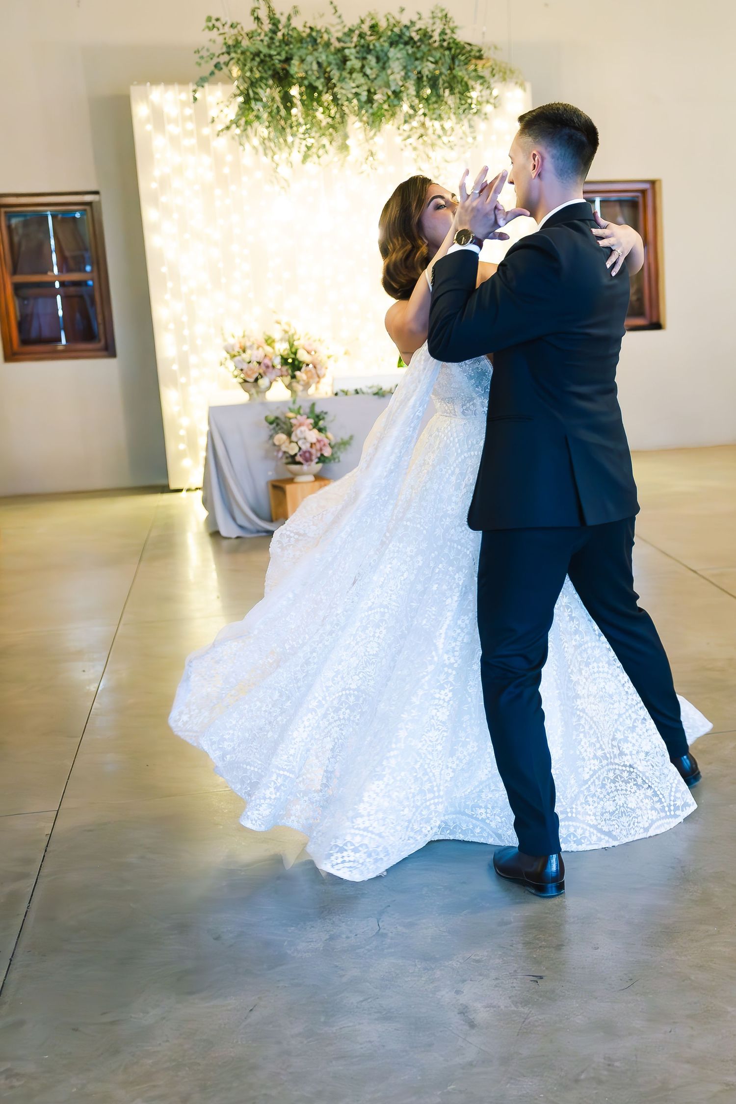 A couple dances gracefully in a ballroom, with the bride's white lace dress flowing elegantly as they twirl.