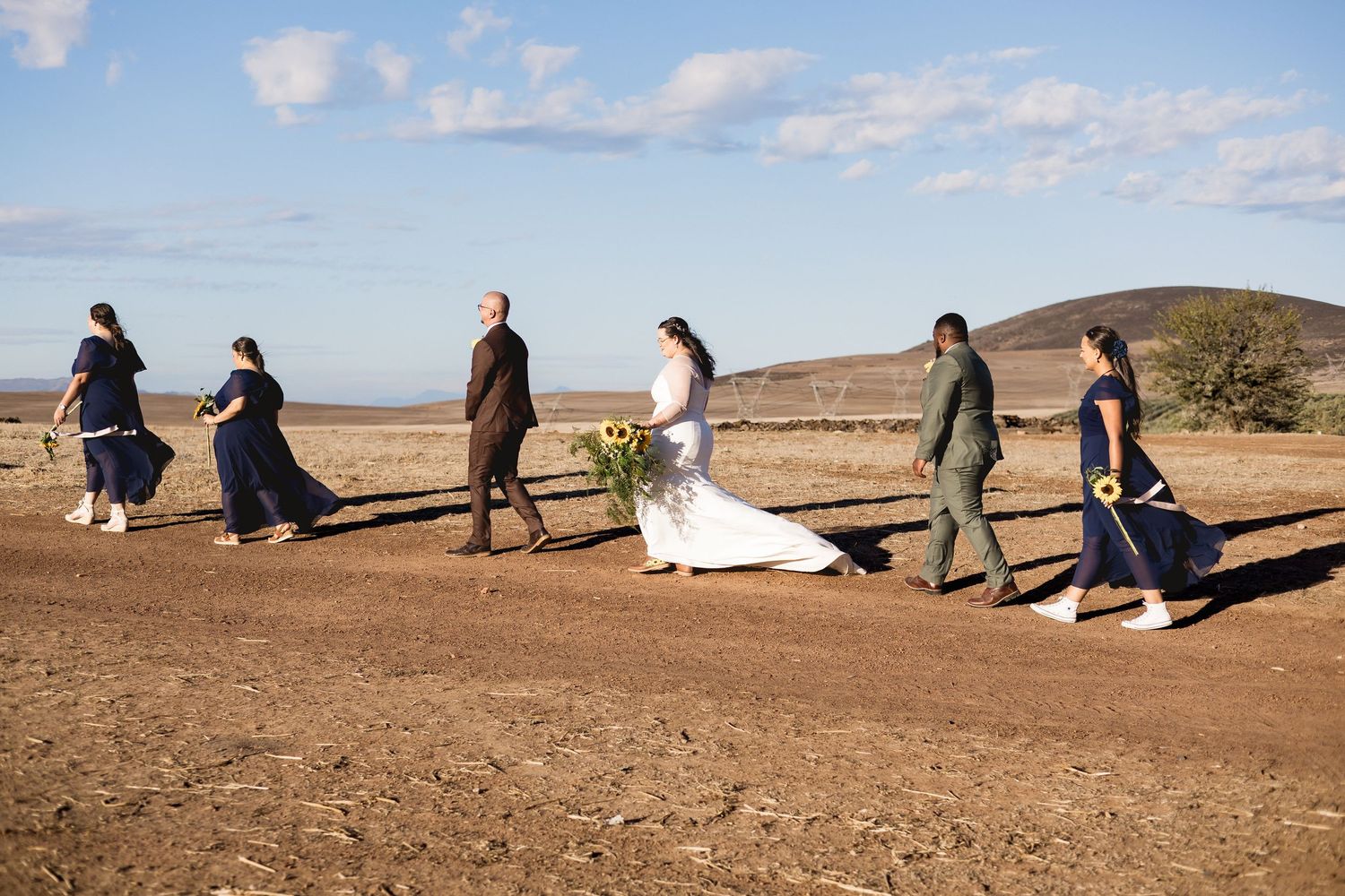 Wedding party walks across rustic dirt path in open landscape, bride in white gown with sunflower bouquet, bridesmaids in navy dresses.