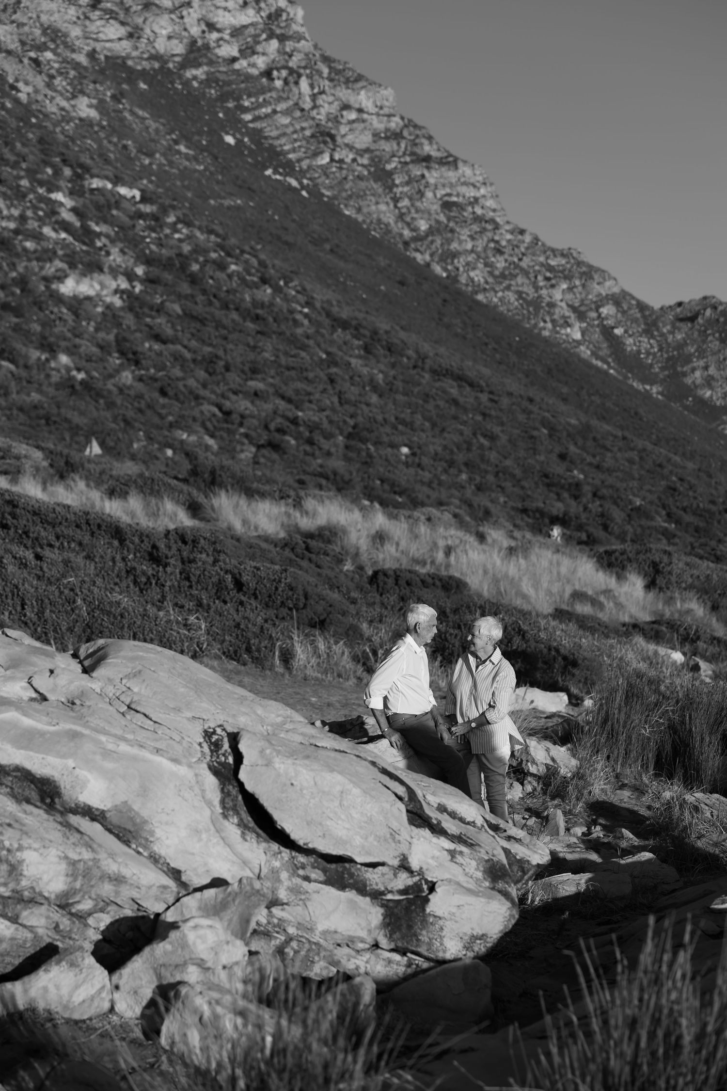 Two elderly people sitting on large rocks with a mountain and lush greenery in the background, black and white photo.
