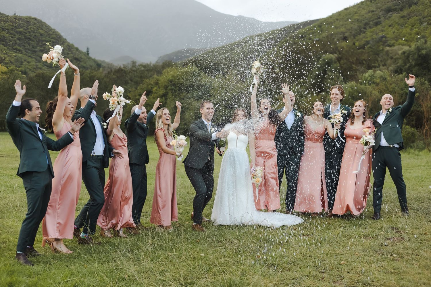 Wedding party members playfully leap and celebrate together against a scenic green mountain landscape in the distance. Popping Champaign.