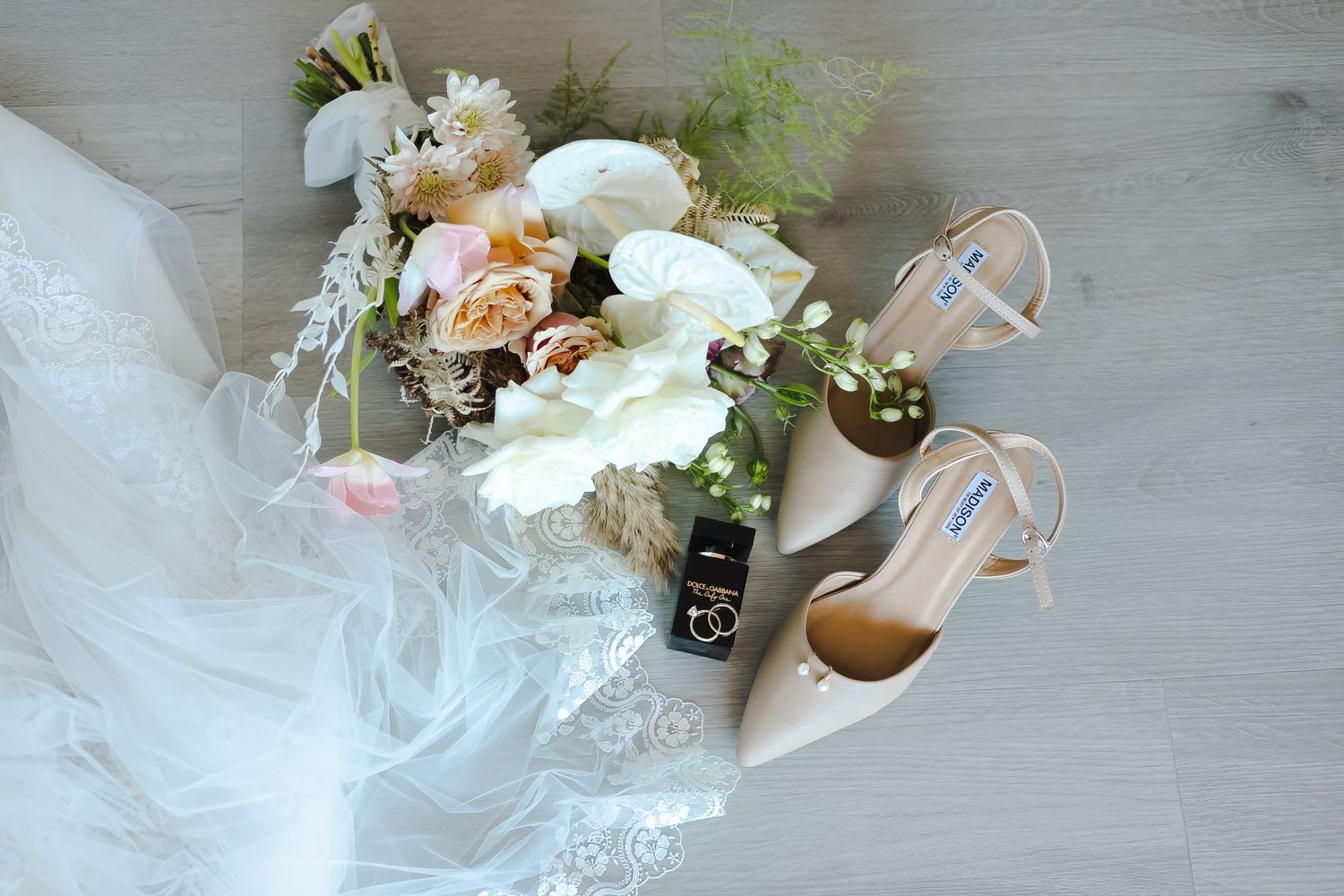 Wedding details including bridal bouquet, shoes and accessories arranged on light wood floor.