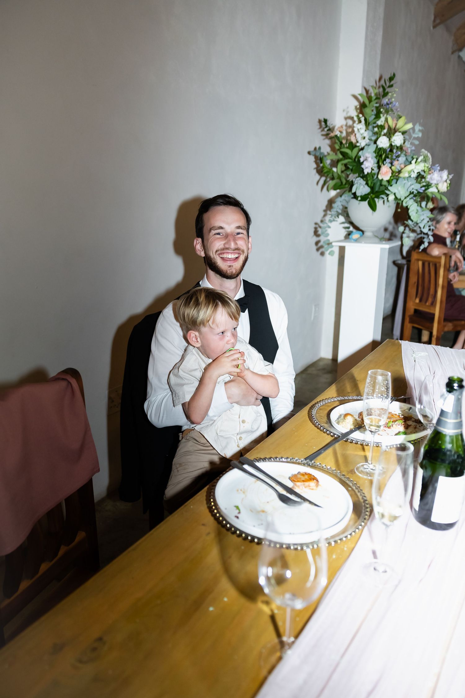 Wedding guests enjoying dinner and conversation at wooden table with white tablecloth and floral centerpiece.