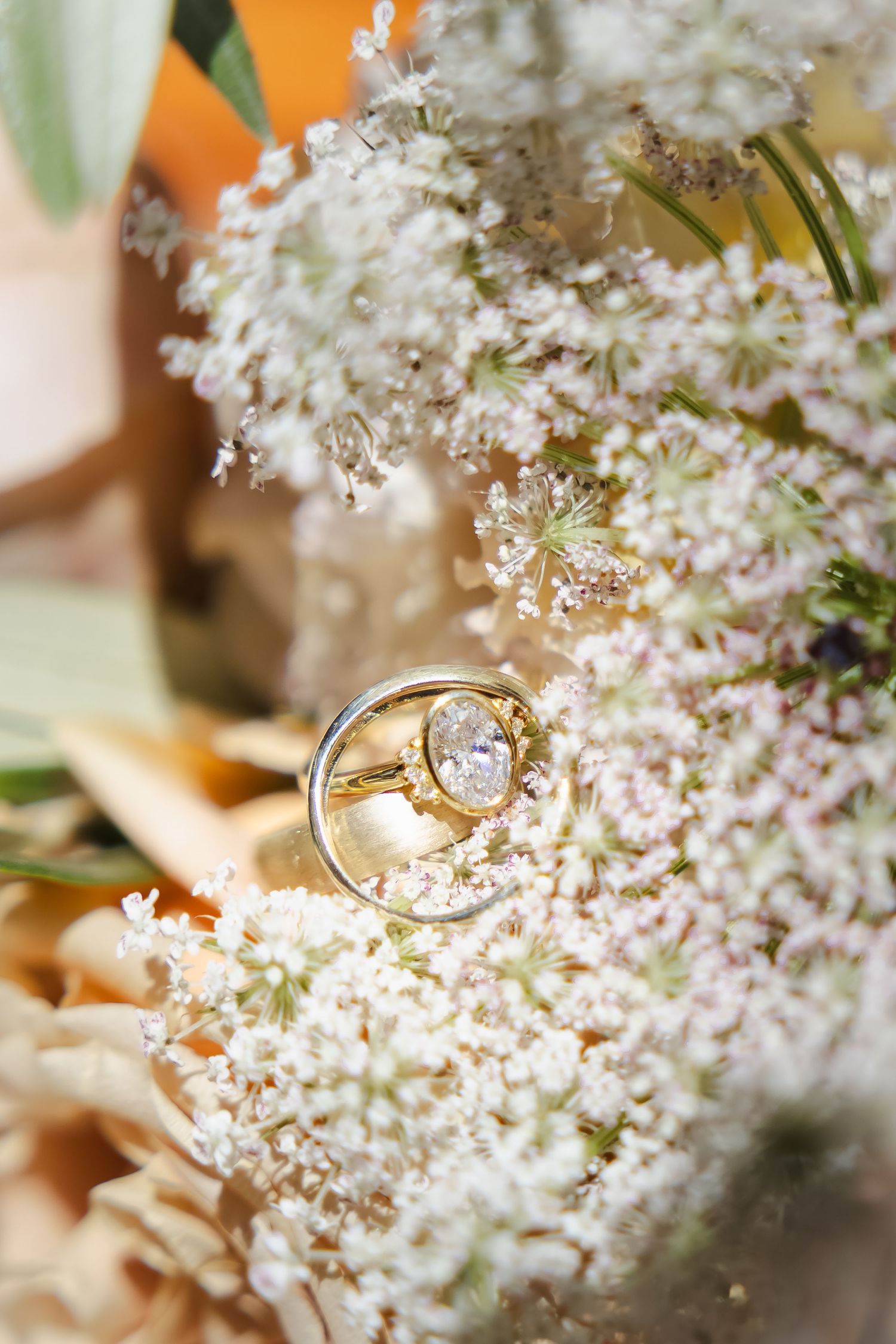 A delicate gold engagement ring with a diamond solitaire nestled among small white Queen Anne's lace flowers.