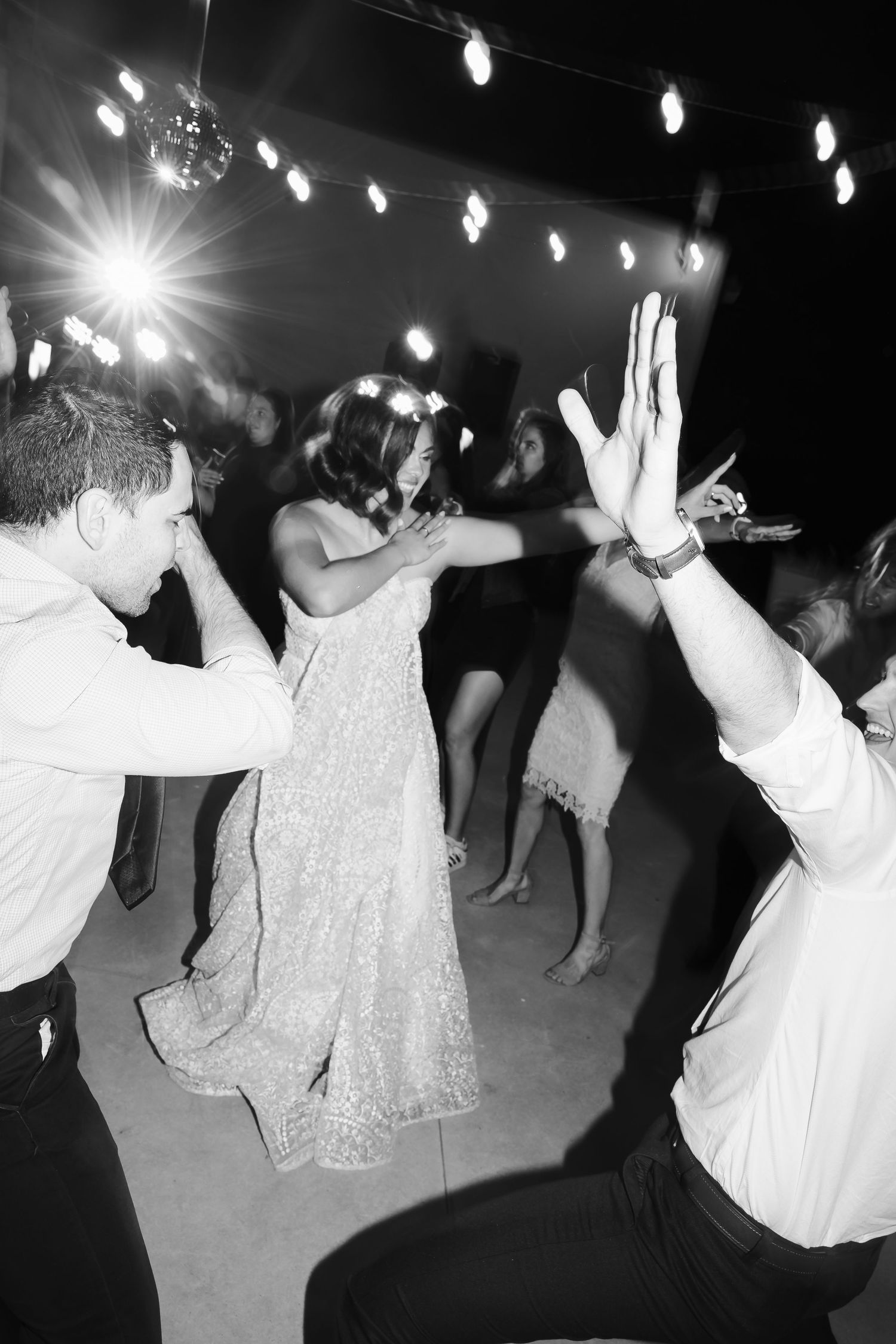 Black and white photo of party guests dancing energetically under string lights at a celebration.