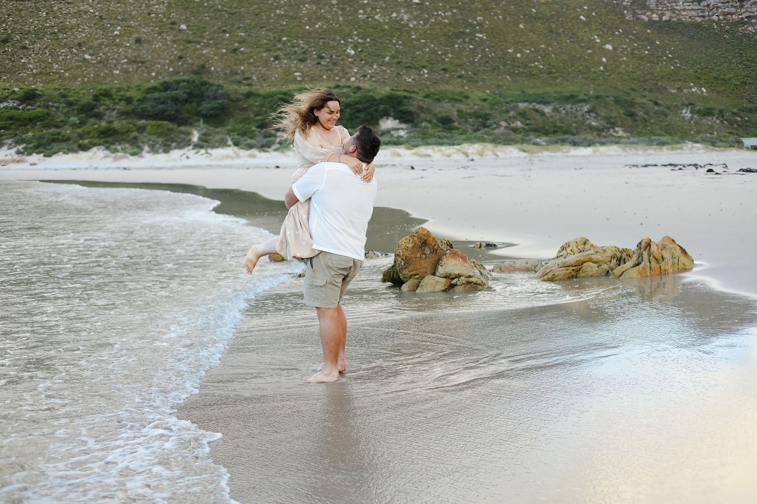 A person in a white dress walks along the shoreline as waves crash against rocks on a scenic beach.