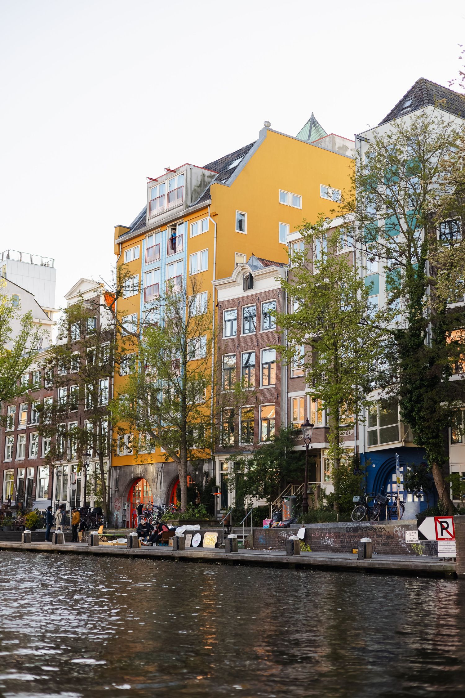 Colorful Amsterdam canal houses with orange facade, trees, and waterfront promenade reflecting in calm canal waters.