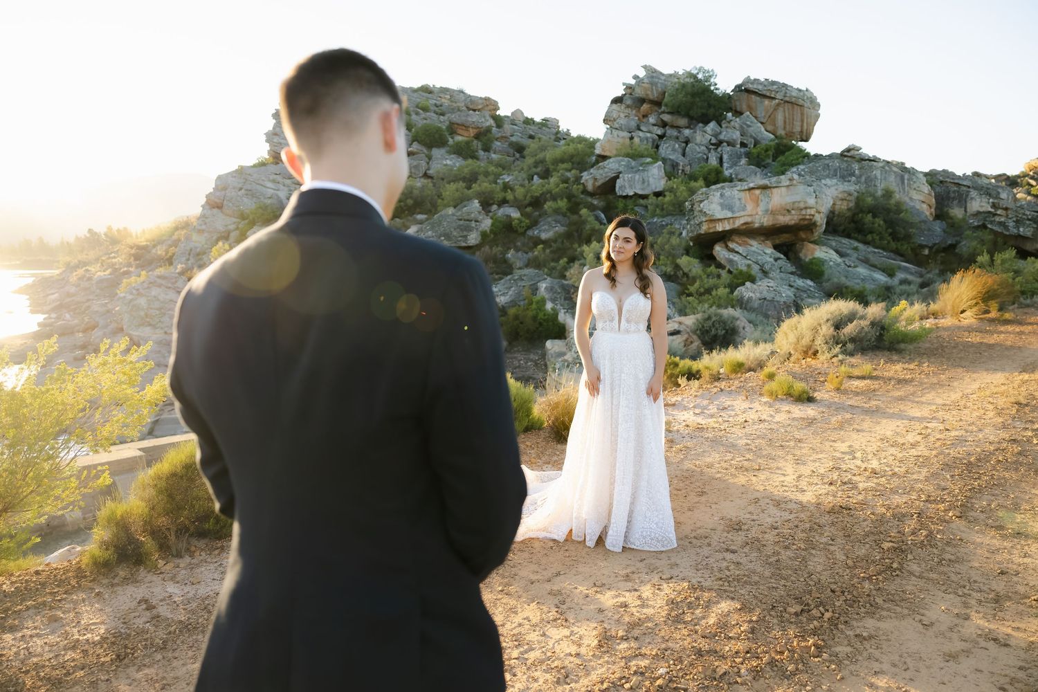 Couple shares romantic moment at sunset on rocky coastal cliff with mountains in background.