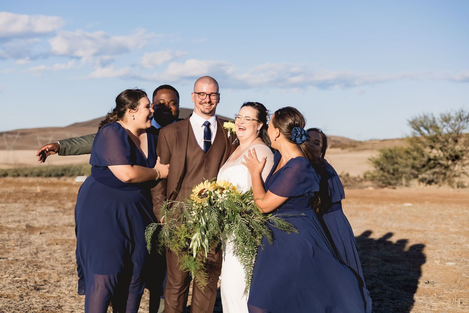 Bride and groom celebrate with bridesmaids in navy dresses outdoors at a rustic desert wedding venue.