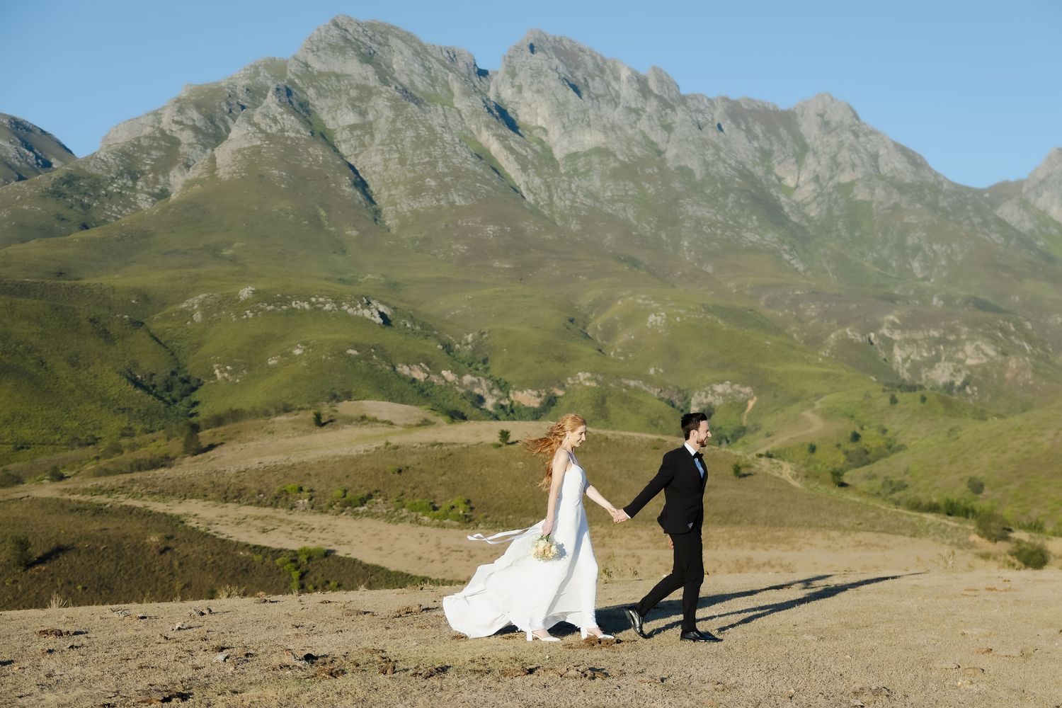 A couple in wedding attire poses against a dramatic mountain landscape backdrop on a winding dirt path.