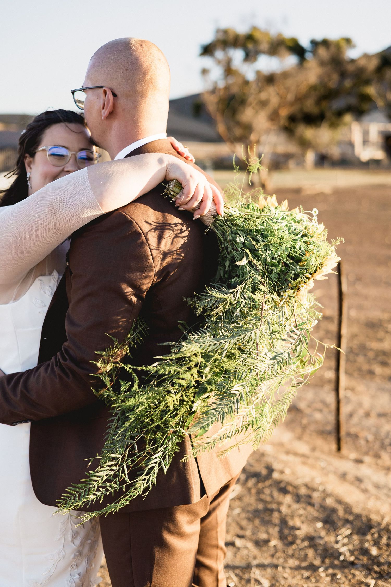 Bride and groom embrace outdoors, she holds a large greenery bouquet, wearing brown suit and white dress.