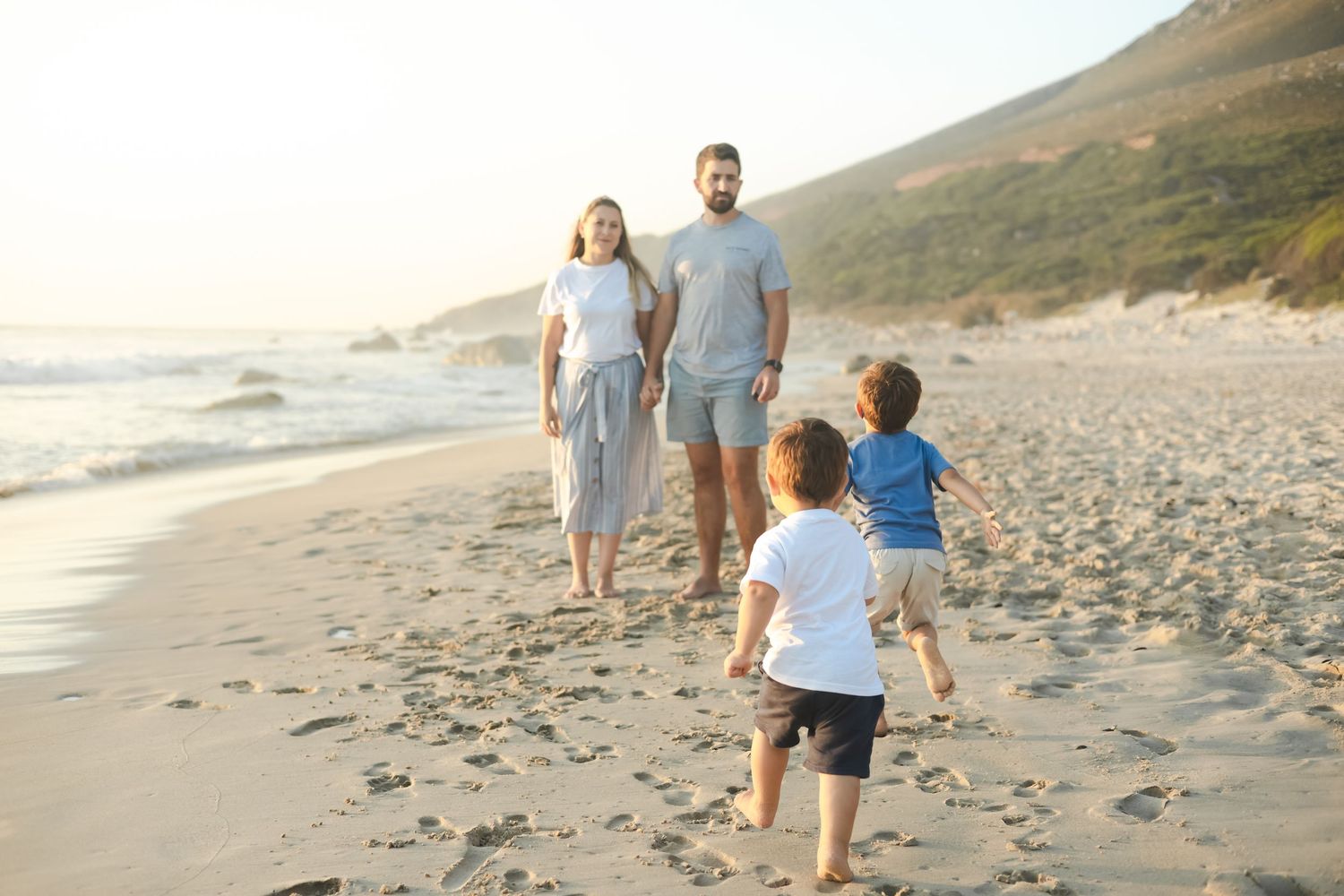 Family of four walking on a sunny beach, parents holding hands while two young children run ahead near the ocean.