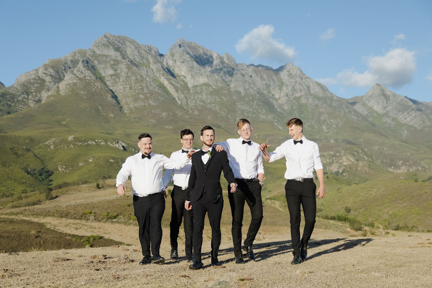 A group of formally dressed people walking along a mountain path with rugged peaks in the background.
