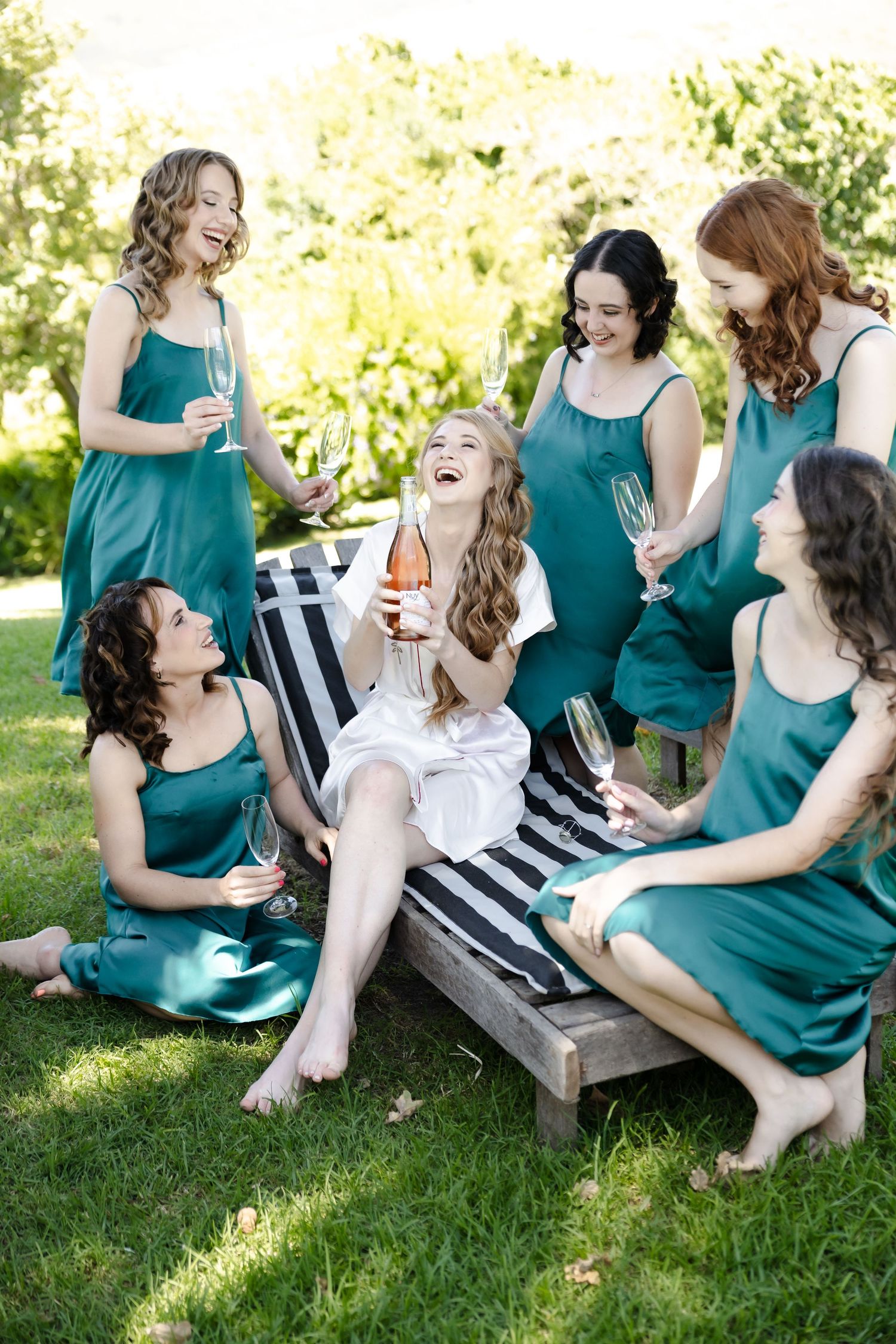 Bridesmaids in teal dresses share joyful laughter while gathered on a garden bench in the sunlight.