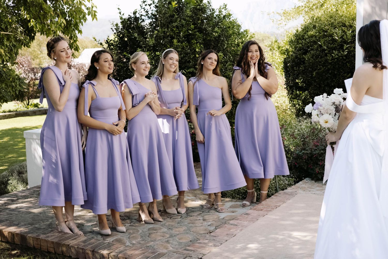 Bridesmaids in lavender dresses sit in a row on garden steps during a wedding photoshoot.