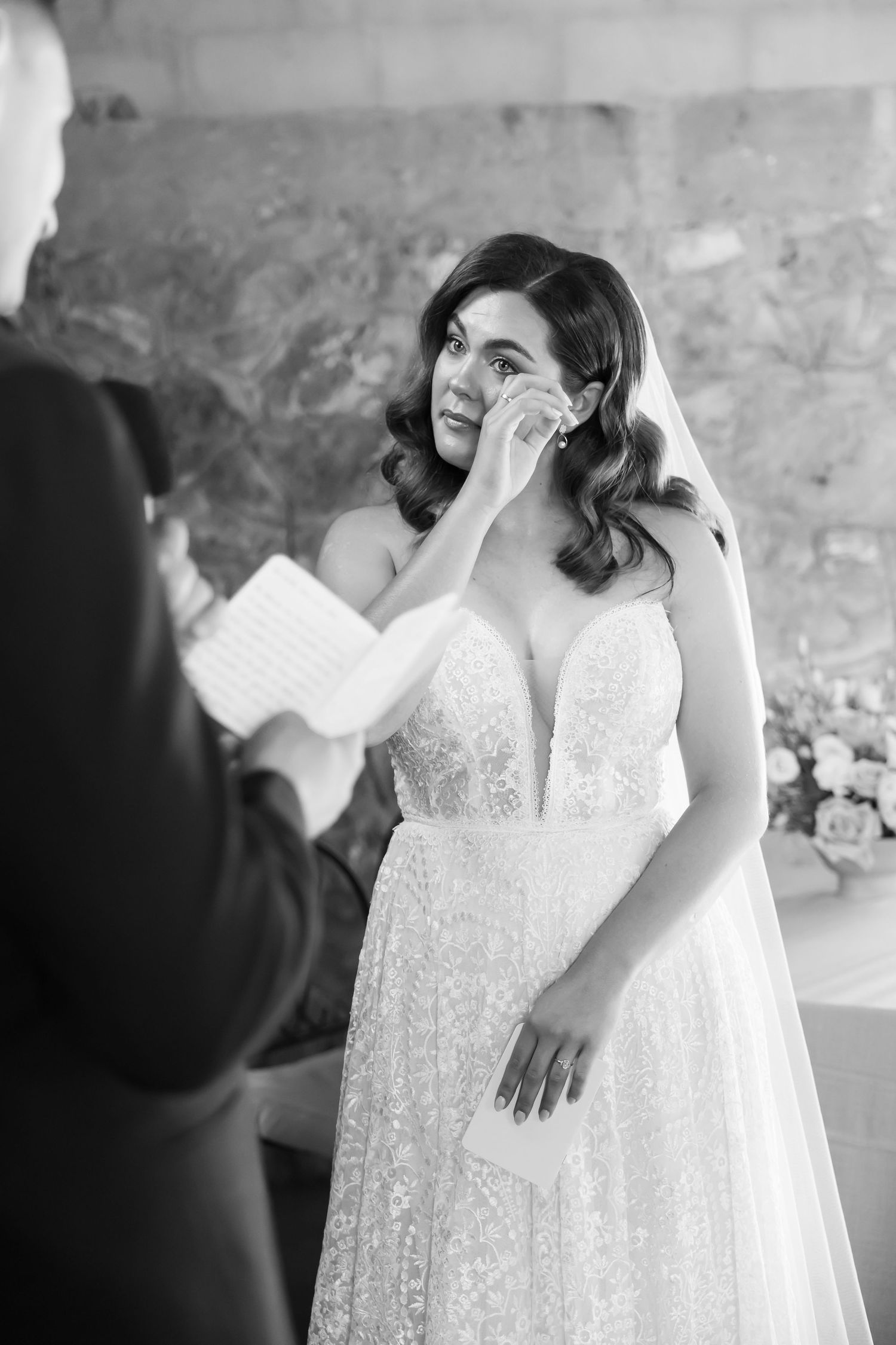 An emotional moment captured in black and white as a bride in a lace wedding dress wipes away tears during the ceremony.