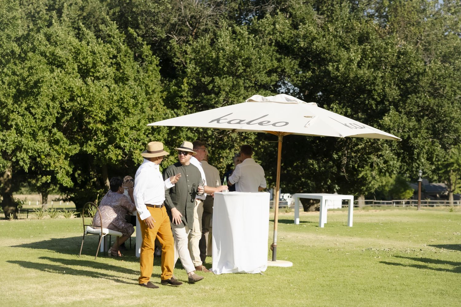People gather at an outdoor event station with white umbrella on a sunny day in a grassy park setting.