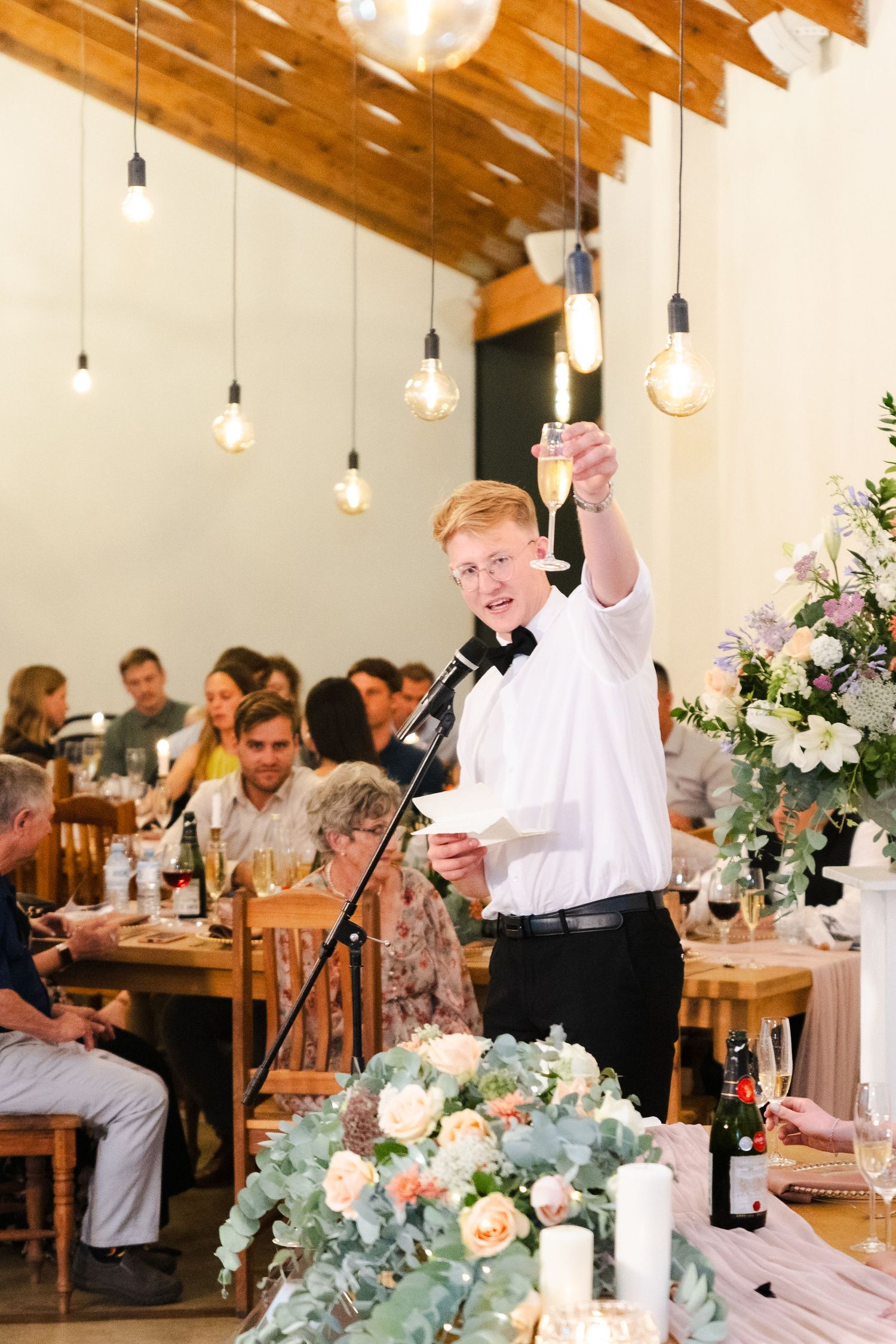 Wedding reception dinner with elegant table settings featuring floral centerpieces and hanging pendant lights in rustic venue.
