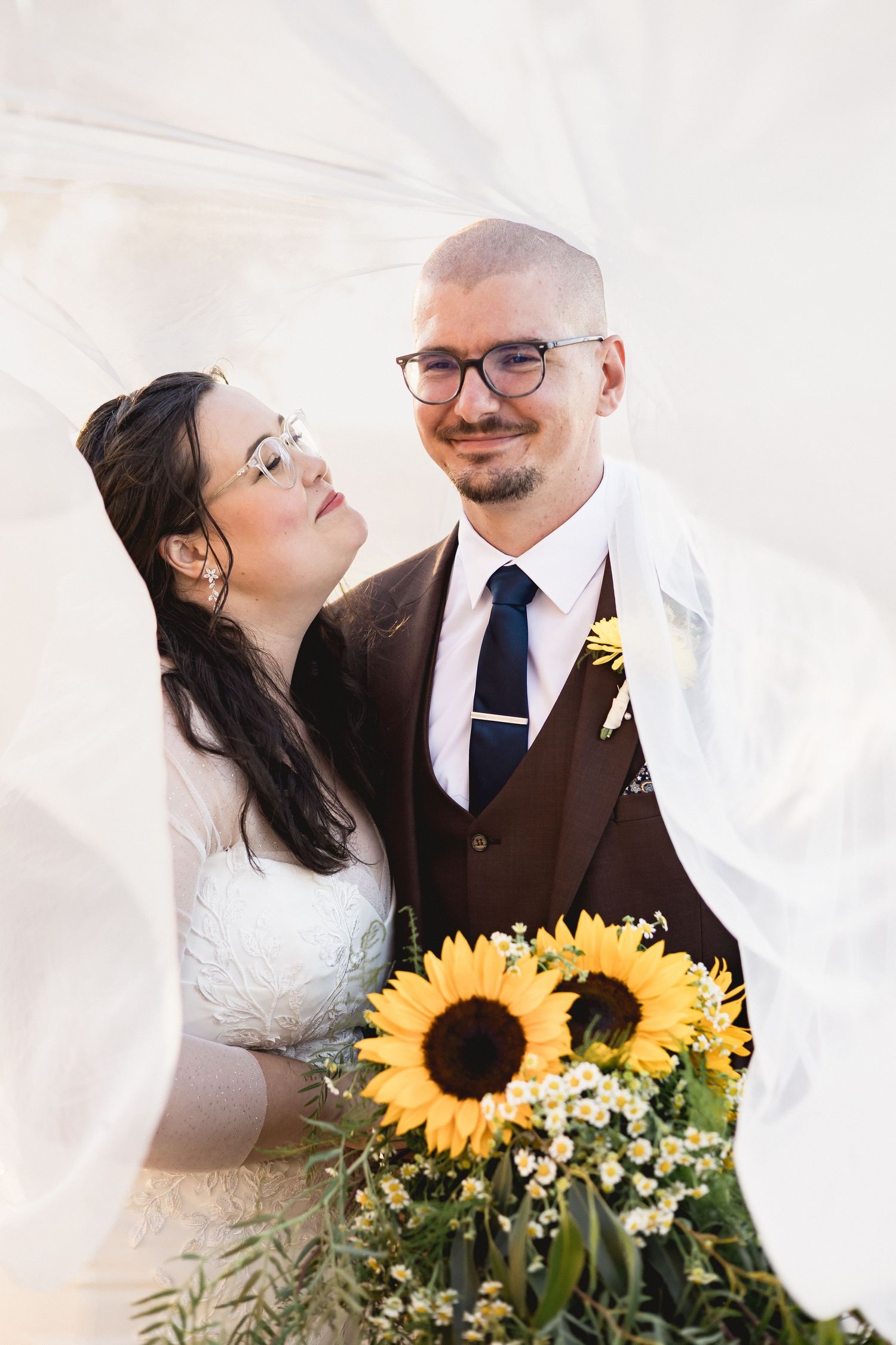 Bride and groom pose under flowing veil holding sunflower bouquet, groom wearing brown suit with navy tie and glasses.