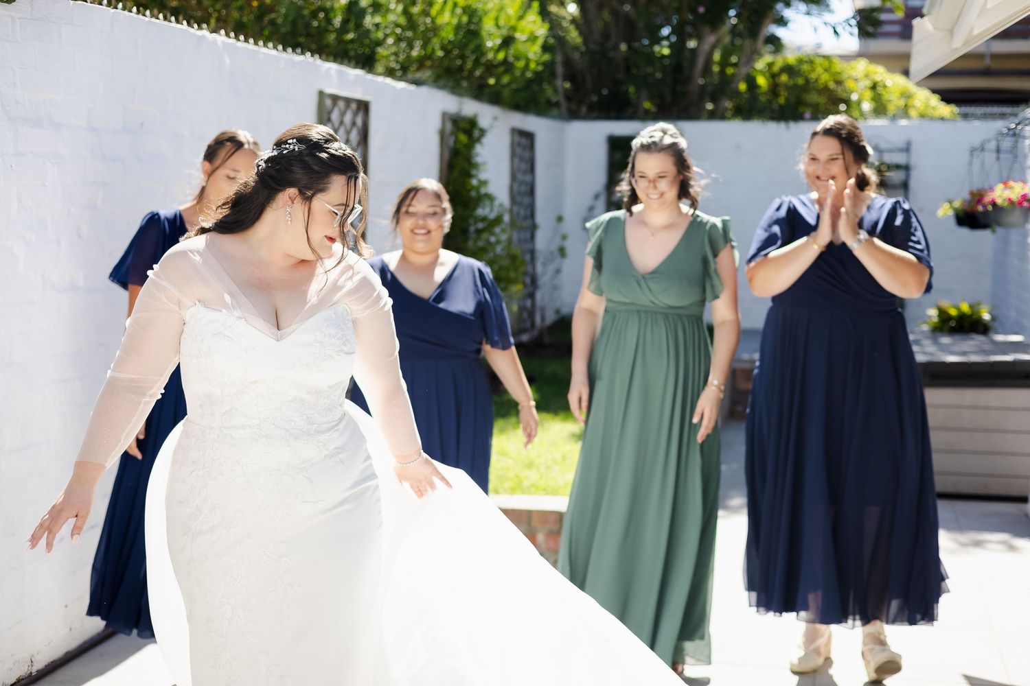 Bride in white gown walks with bridesmaids in navy and green dresses outdoors on sunny wedding day.