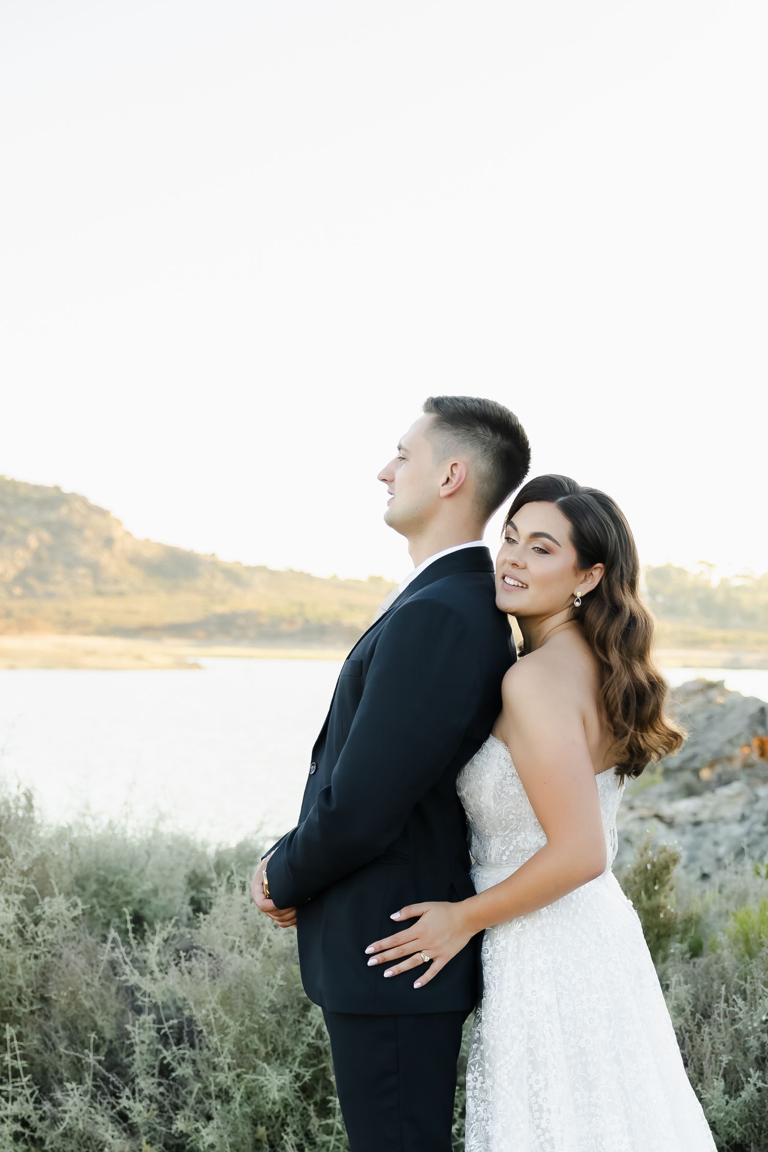 Newlyweds share an intimate moment against a serene waterfront backdrop at sunset.