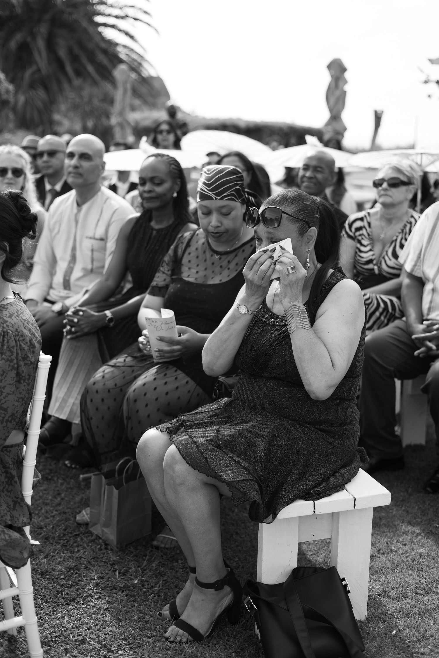 Mourners in black attire sit solemnly on white chairs at an outdoor memorial service.