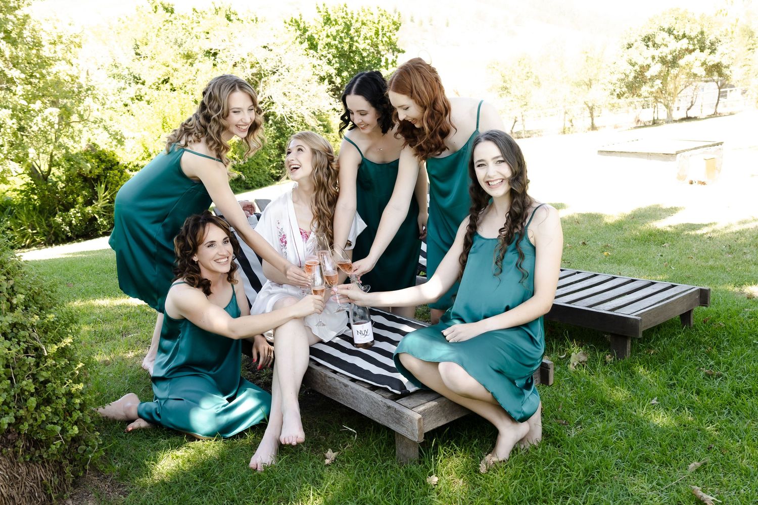 A group in teal dresses sits together on a wooden bench in a sunny outdoor garden setting.