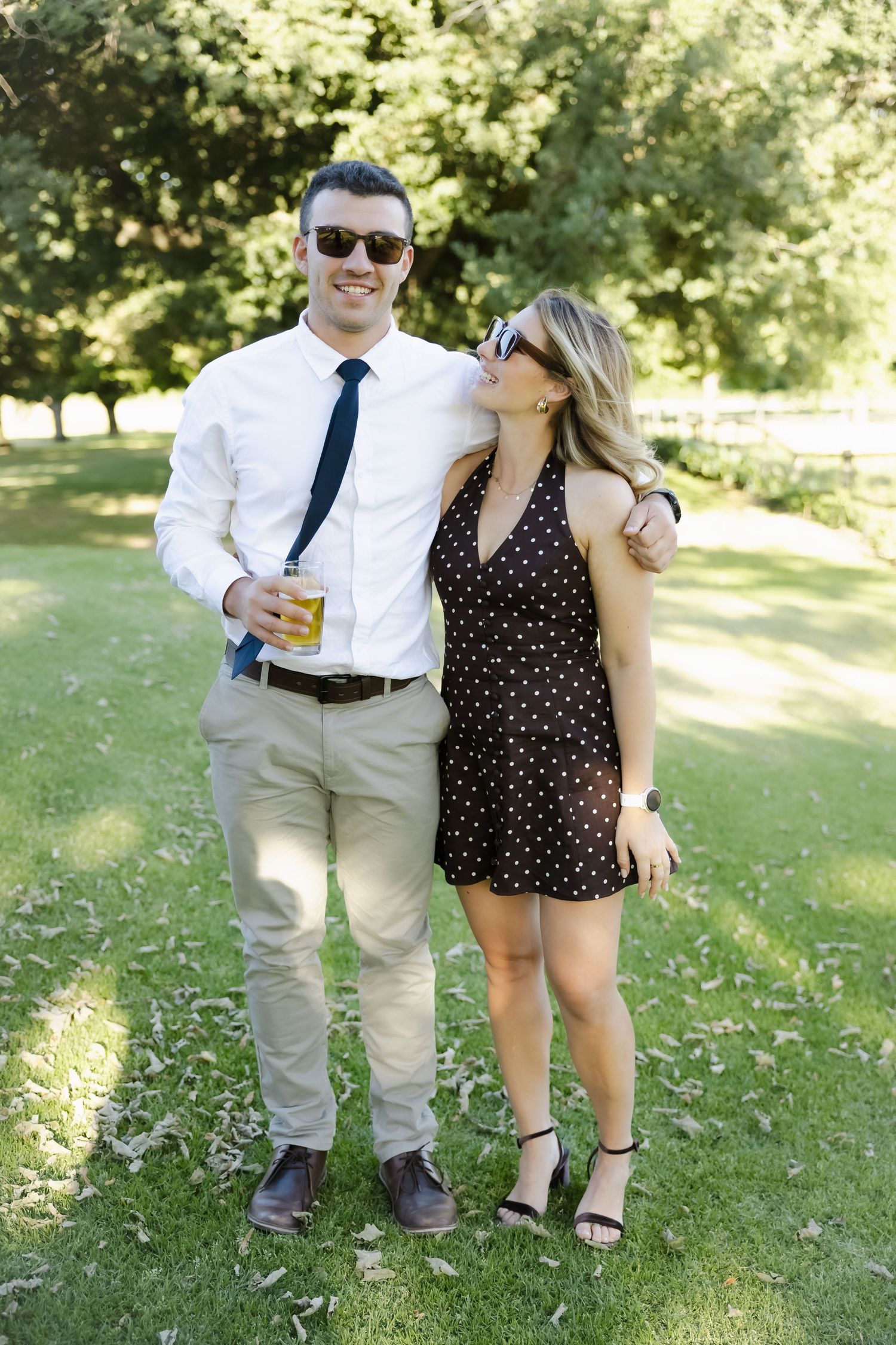 A couple in formal attire shares a playful moment together on a sunny lawn during a summer outdoor event.