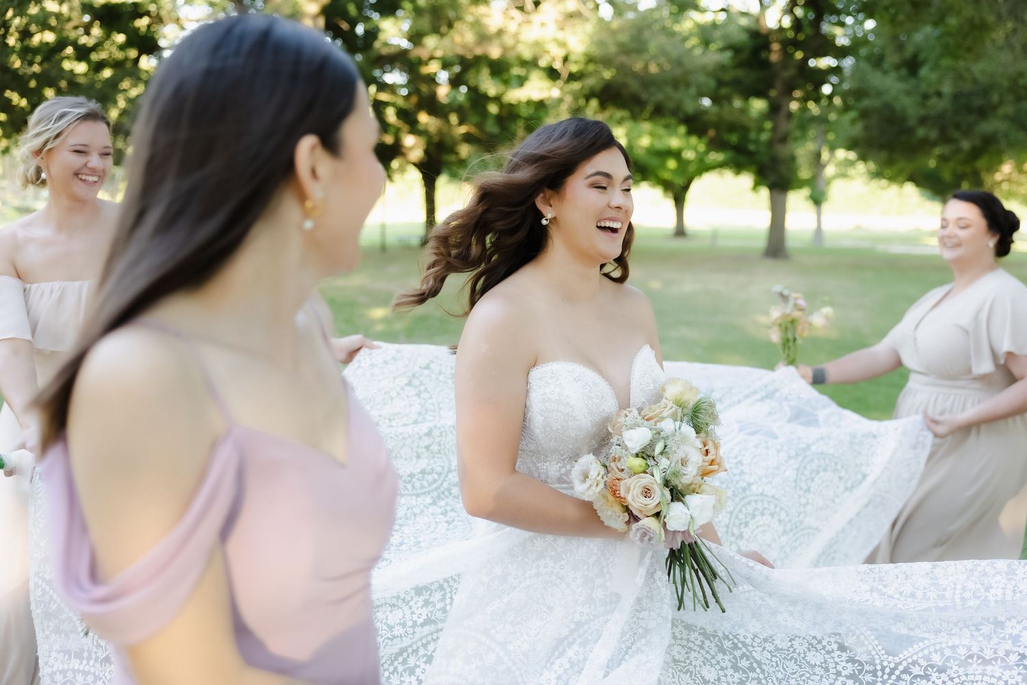 A group of people in white dresses laugh together while holding bouquets in an outdoor garden setting.