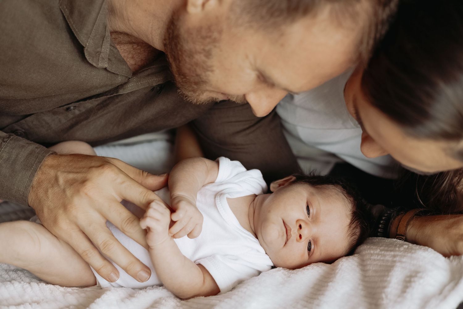 Parents sharing an intimate moment with their baby on white bedding.