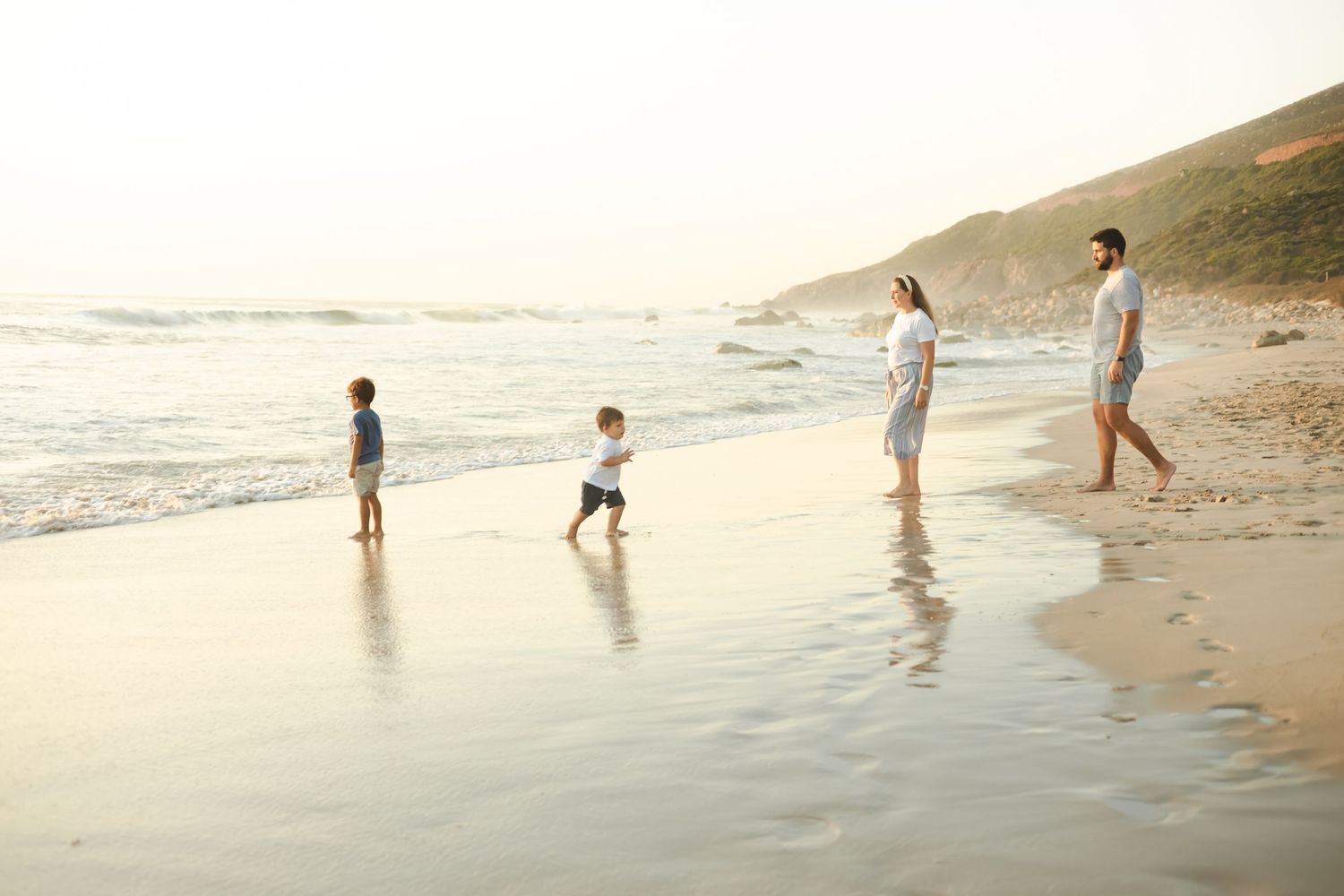 Family of four enjoying a sunny beach walk along the shoreline with waves and sandy hills in the background.