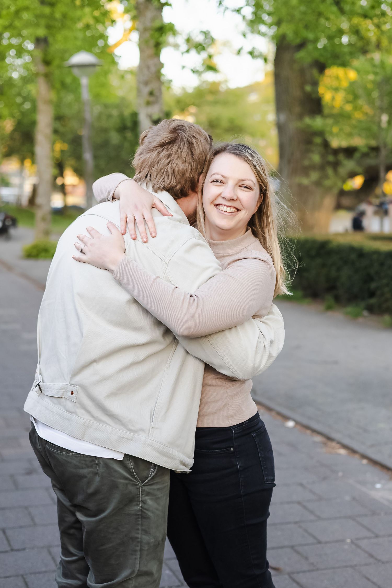 A smiling blonde woman hugging a man outdoors on a tree-lined brick pathway on a sunny day.