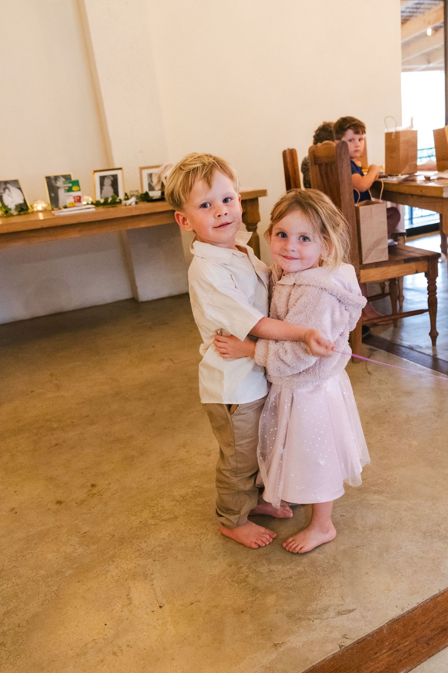 Two young toddlers in casual clothing hug and dance together on a concrete floor.