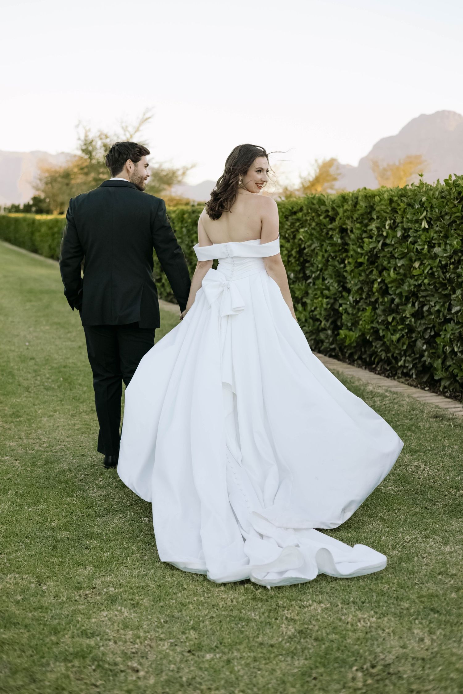 Wedding couple walking across a manicured lawn with hedge backdrop.