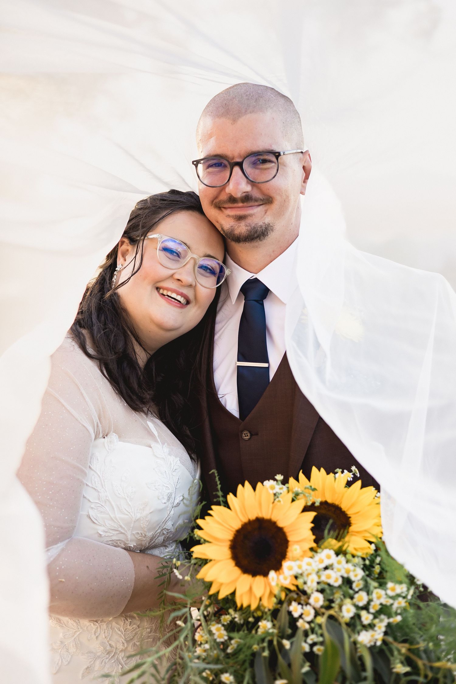 Happy couple in glasses smiling under flowing veil, groom in brown vest holding sunflower wedding bouquet.