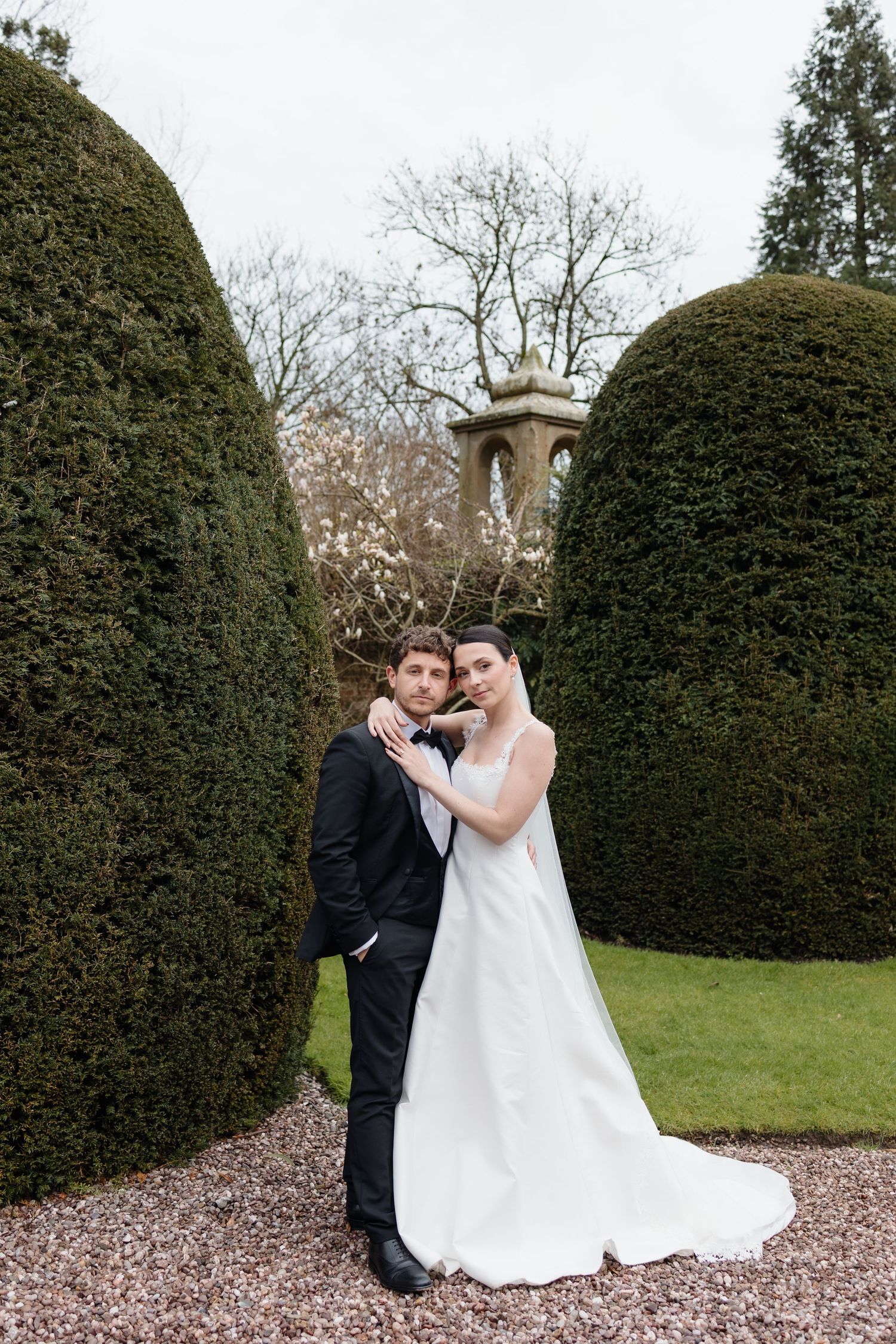 Bride and groom on the grounds of Soughton Hall. Stood in front of topiary bushes, embracing