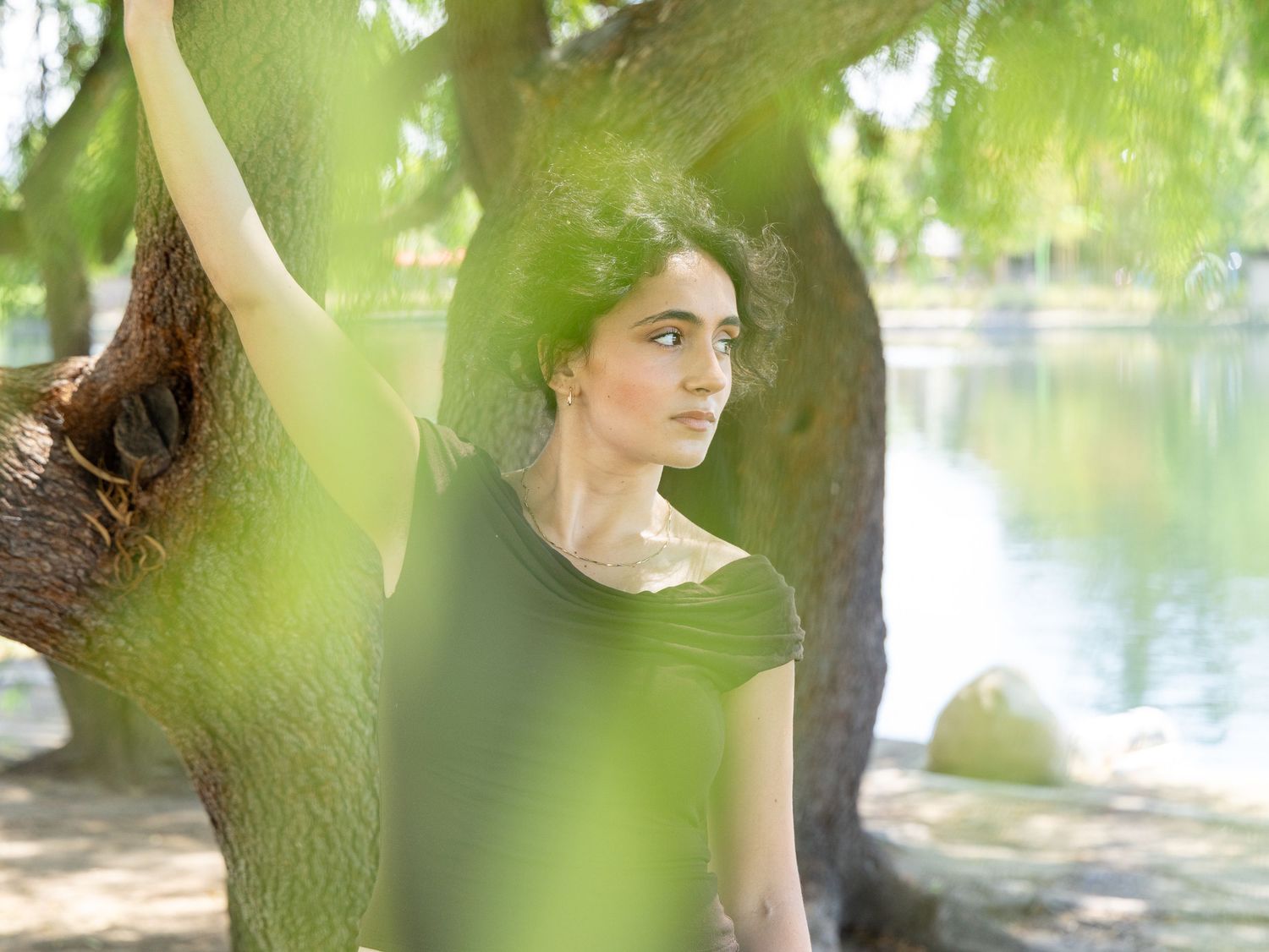 Young woman in green top leaning against a tree by a serene lake in a lush park setting.