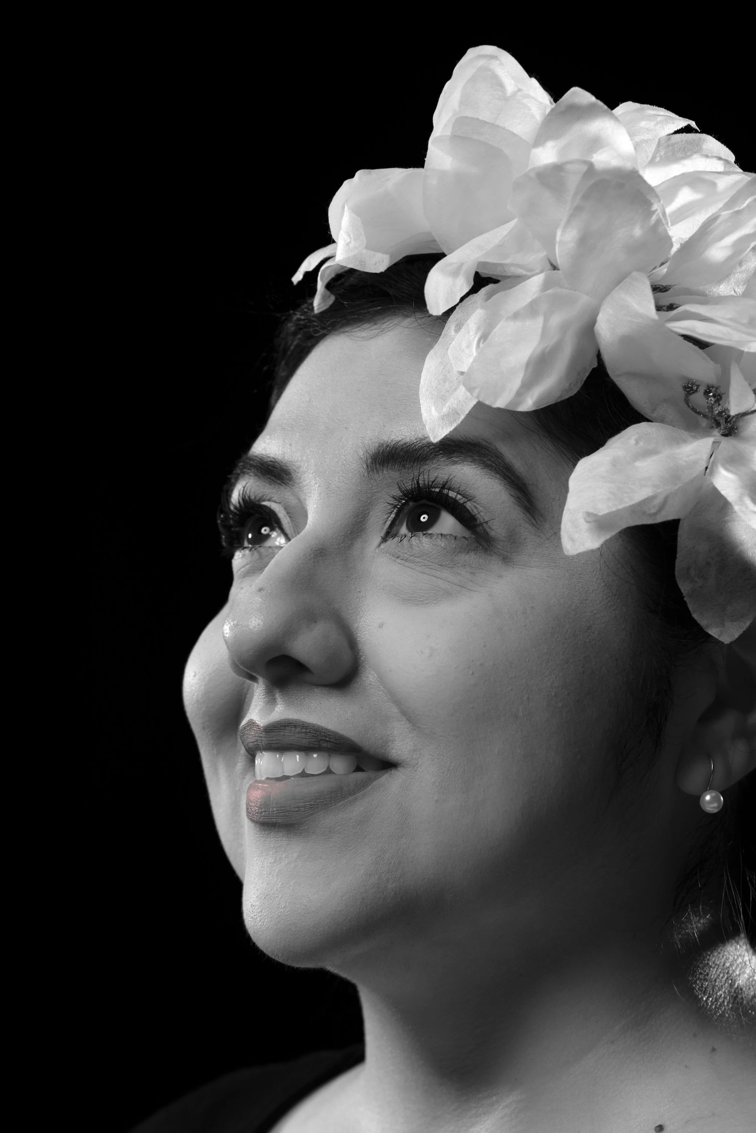Black and white portrait of a smiling woman wearing large white flowers in her hair against a dark background.