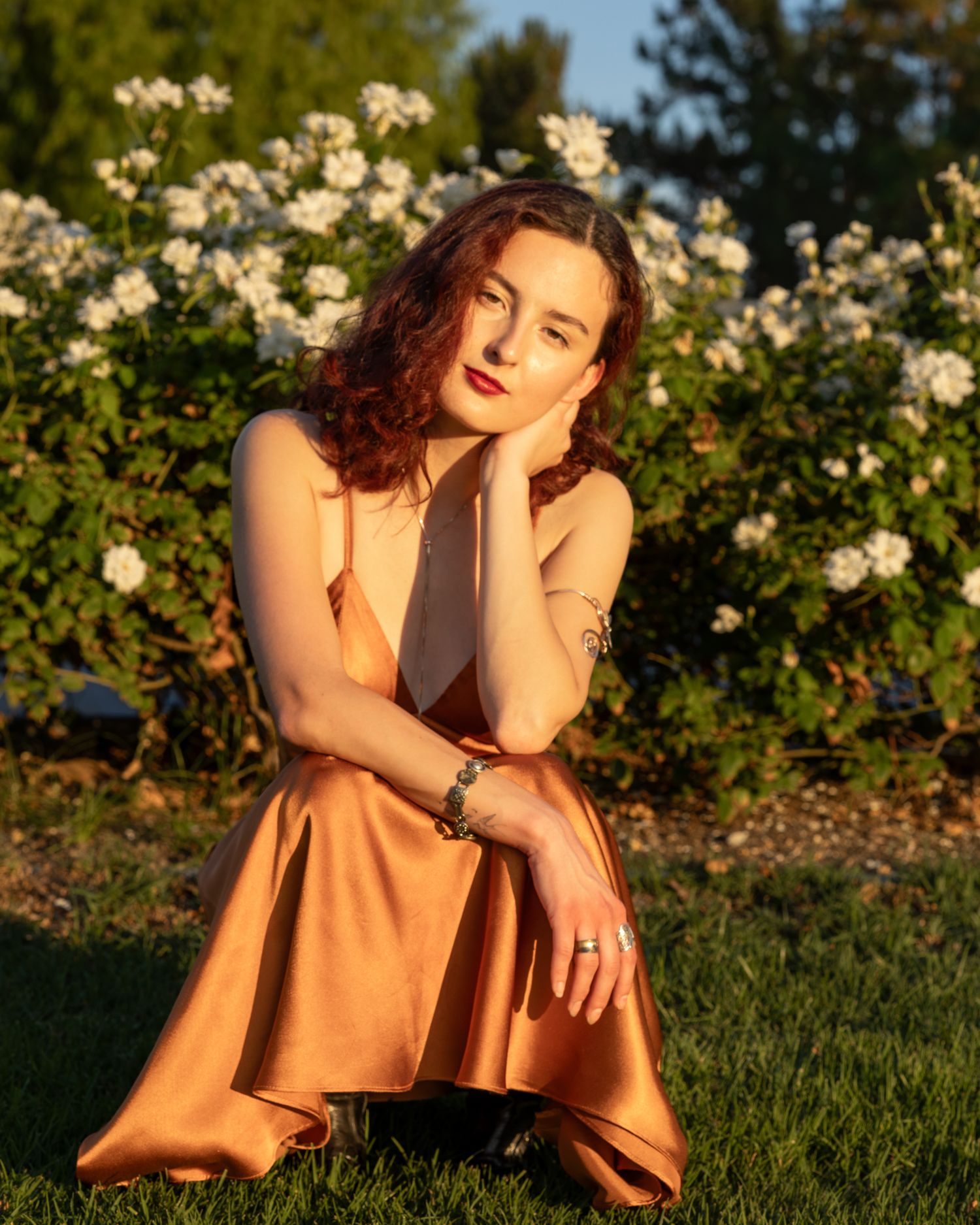Young woman in copper satin dress poses gracefully outdoors near white roses in golden hour sunlight.