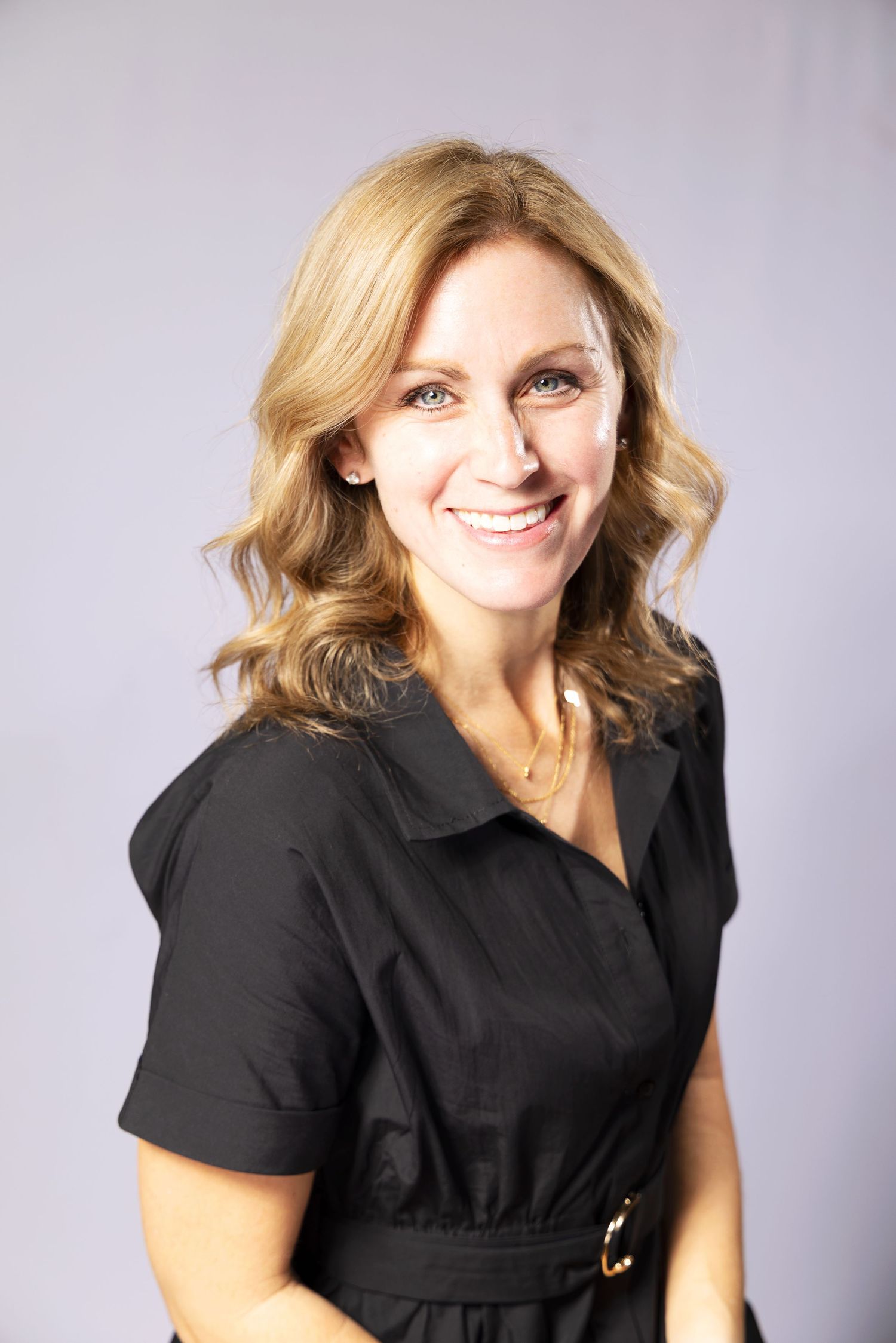 Professional portrait of a smiling blonde woman in a black dress against a light gray studio background.