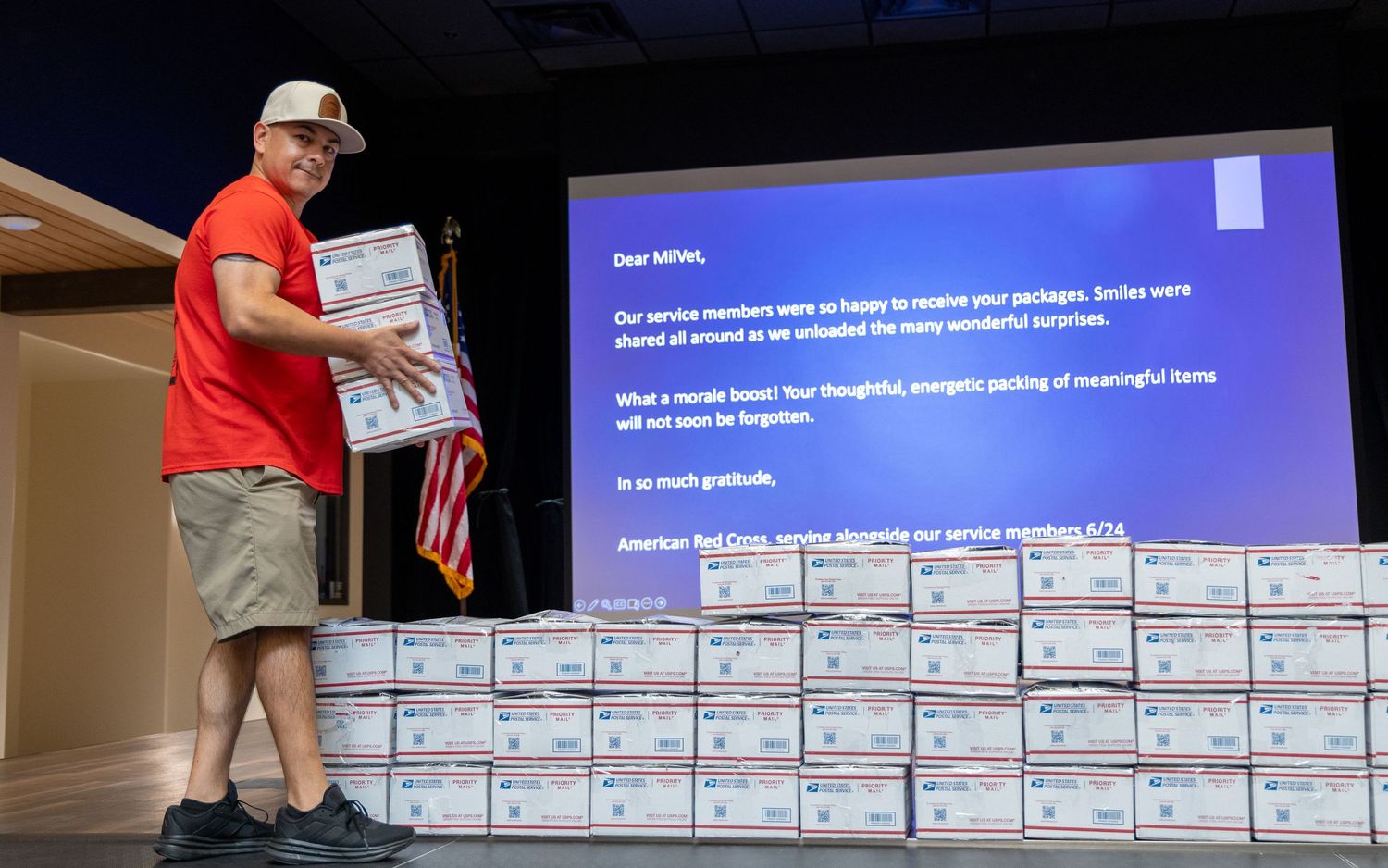 Man in red shirt stands beside stacked care packages with American flag, Red Cross volunteer appreciation event.