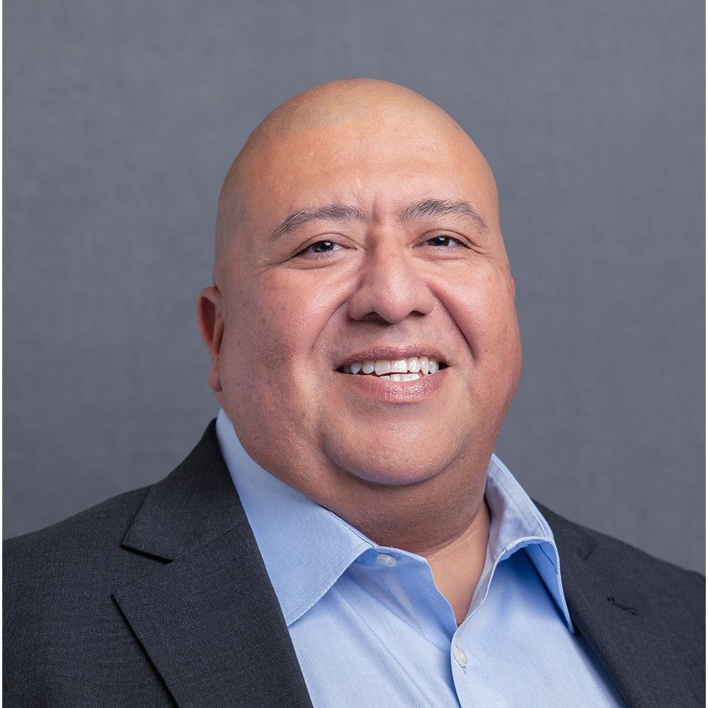 Professional headshot of a bald man in a dark blazer and light blue shirt smiling against a gray background.