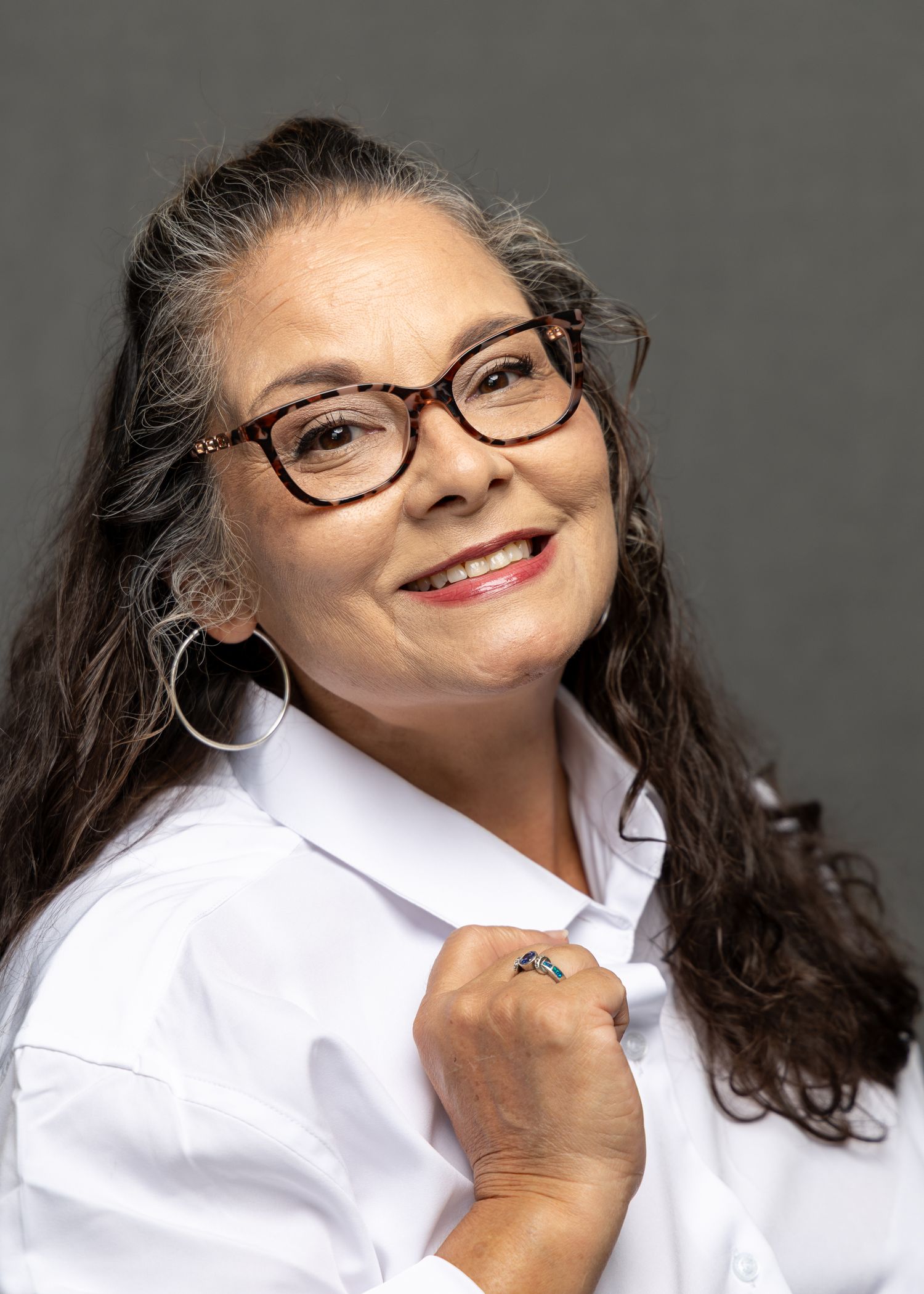 Smiling middle-aged woman with glasses, gray-streaked dark hair, hoop earrings, and white shirt poses against gray background.